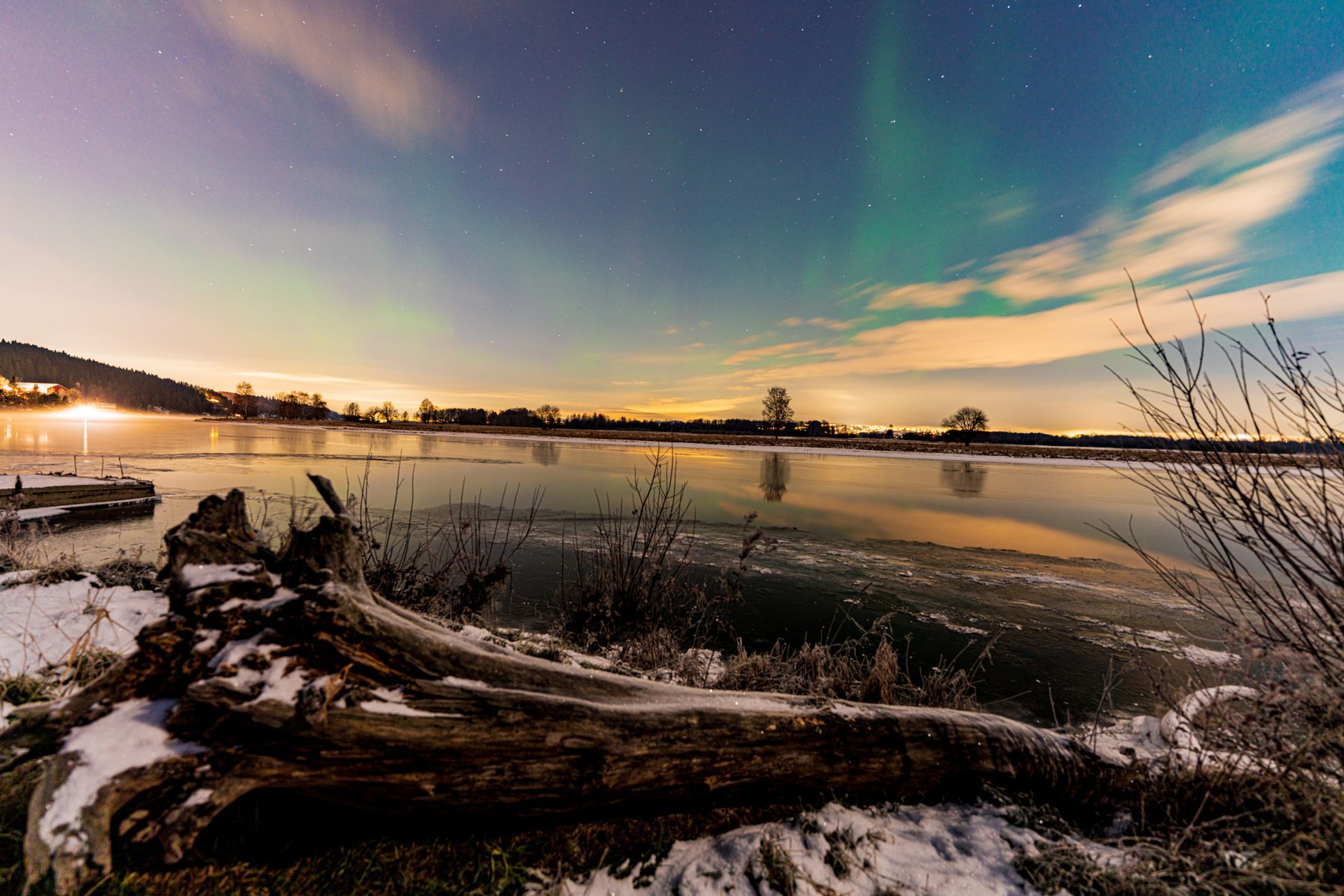 Het noorderlicht verlicht de nachtelijke hemel boven een met sneeuw bedekt, ijzig landschap met een grote boomstam op de voorgrond.