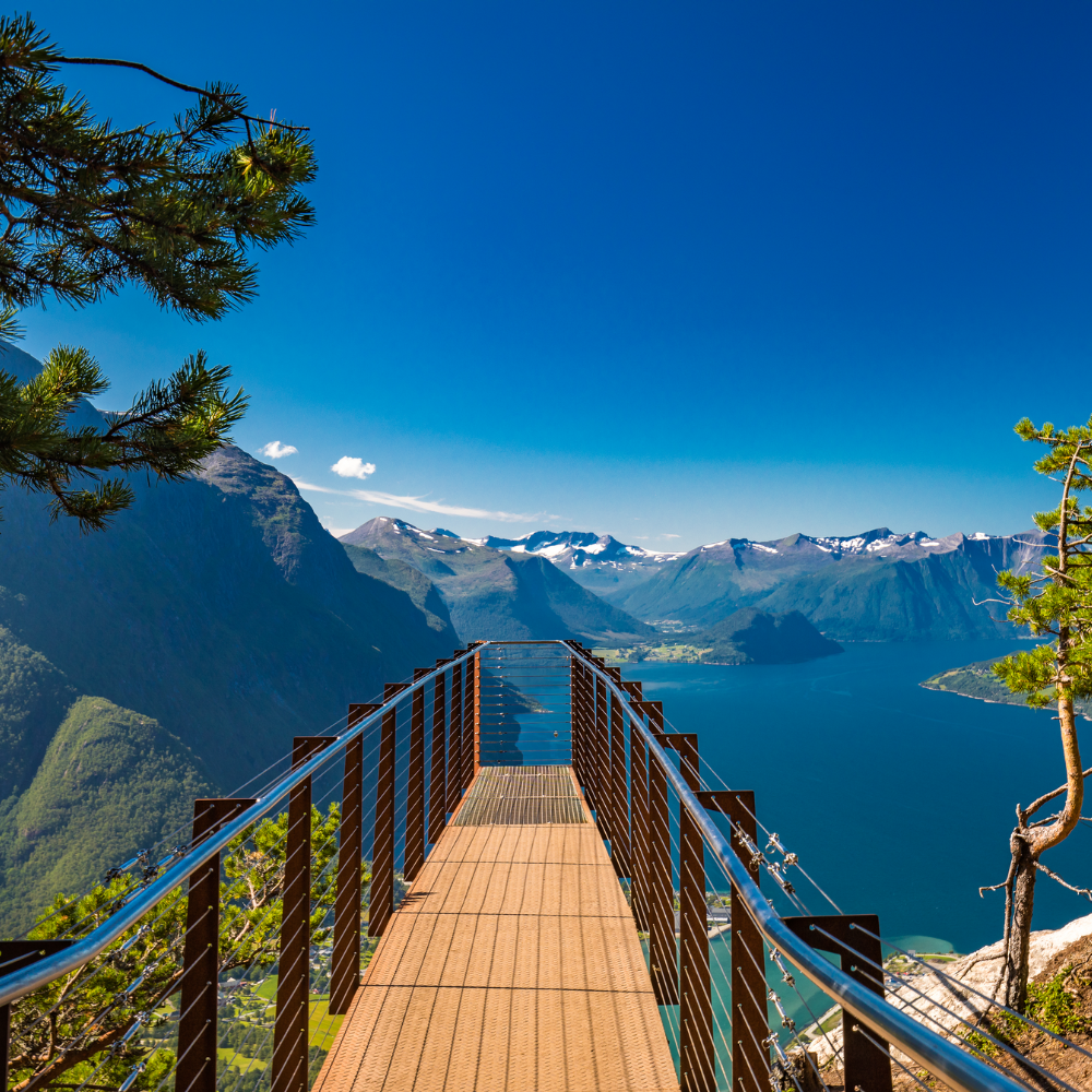 Een houten uitkijkplatform strekt zich uit over een steile klif en biedt uitzicht op een diepblauwe fjord, omgeven door torenhoge bergen.