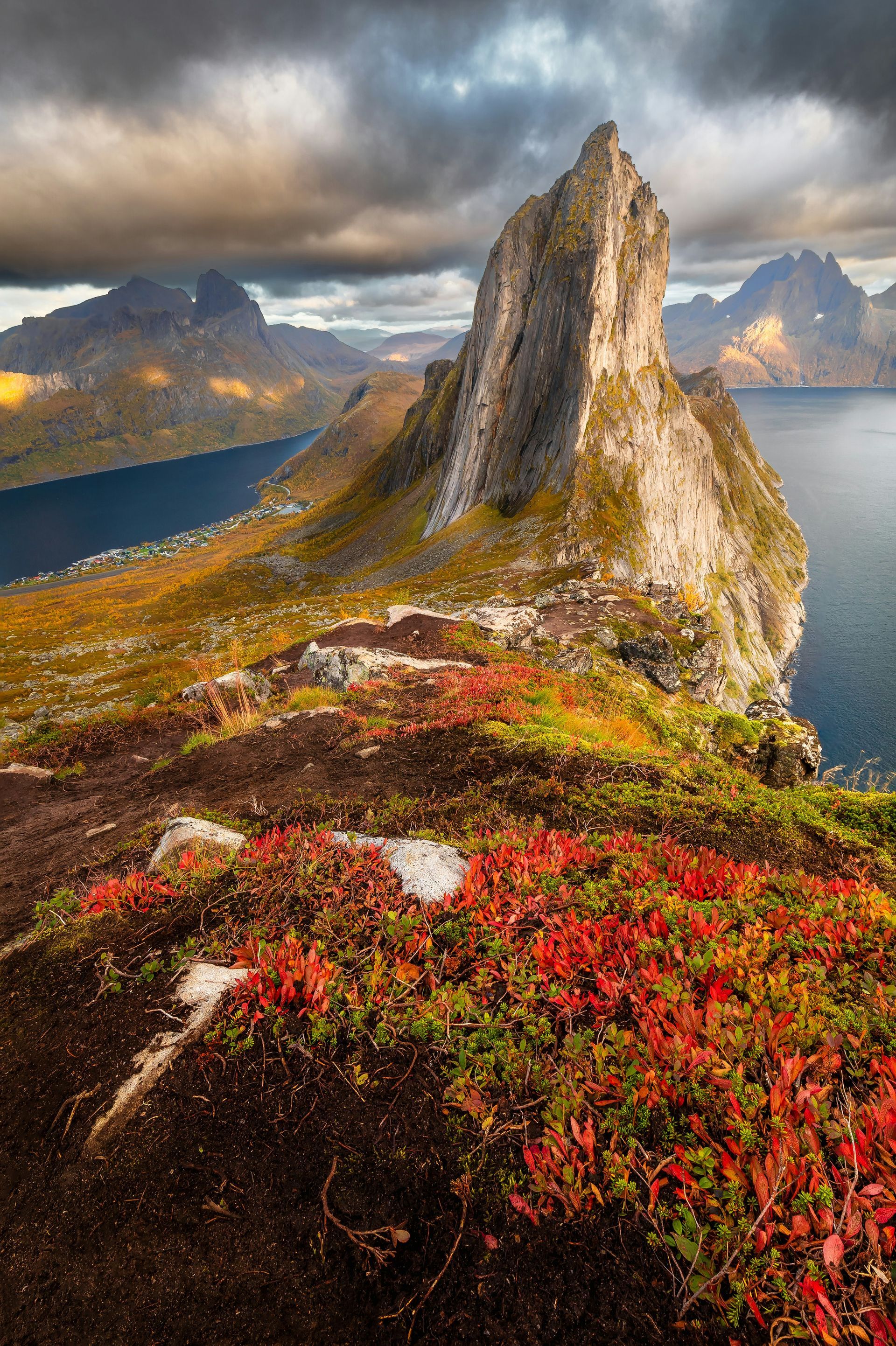 Een torenhoge, grillige rotspiek rijst op boven een diepblauwe fjord, omgeven door levendig herfstblad onder een stormachtige hemel.