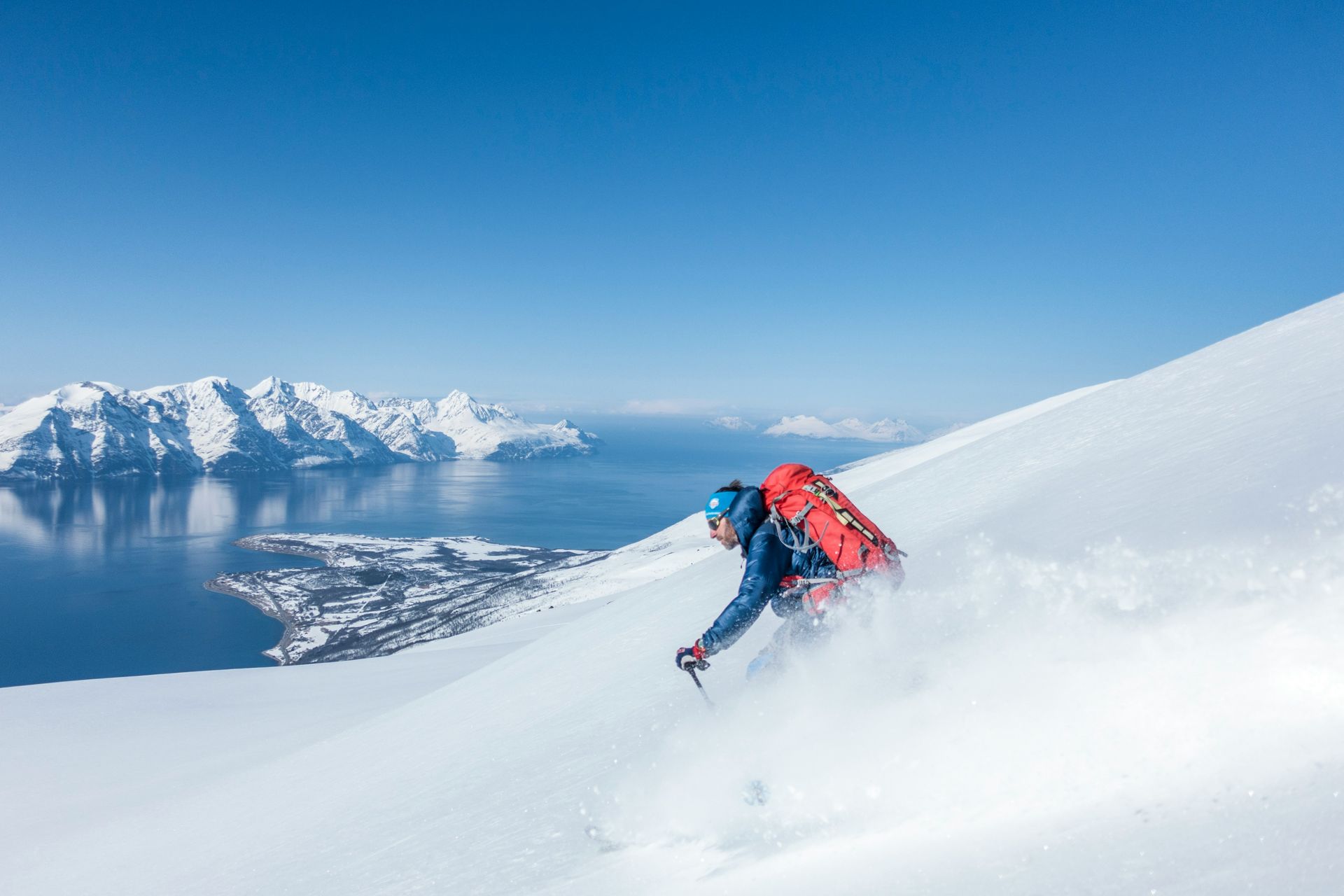 Een persoon met een rugzak is aan het skiën op een met sneeuw bedekte berg met uitzicht op het water.
