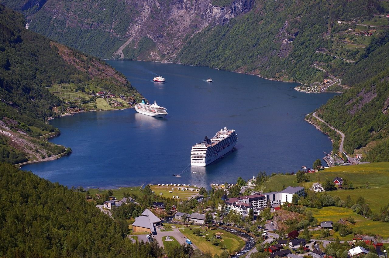 Twee grote cruiseschepen in een diepblauwe Noorse fjord, omgeven door weelderig bergachtig terrein en een klein dorpje.