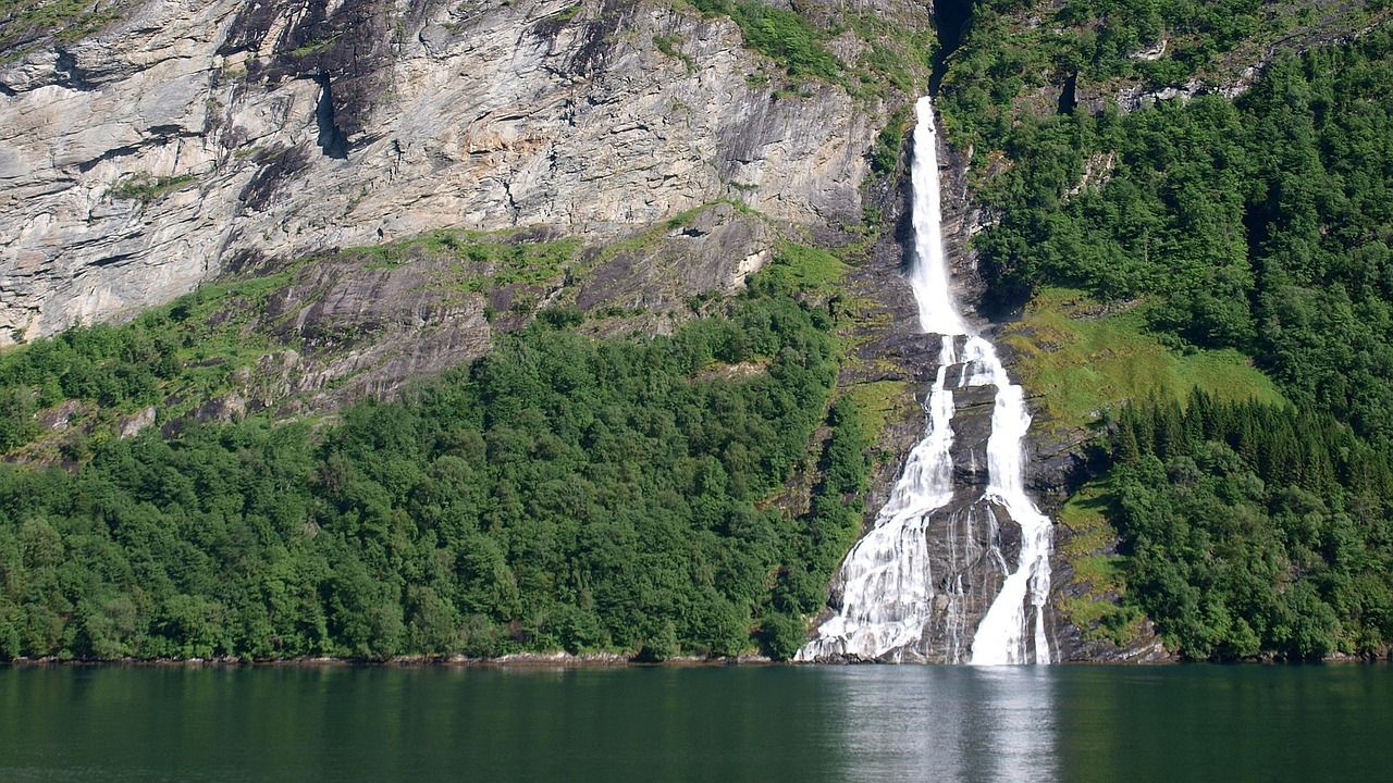 Een waterval stroomt van een klif naar beneden en komt in een fjord terecht dat omgeven is door bomen.