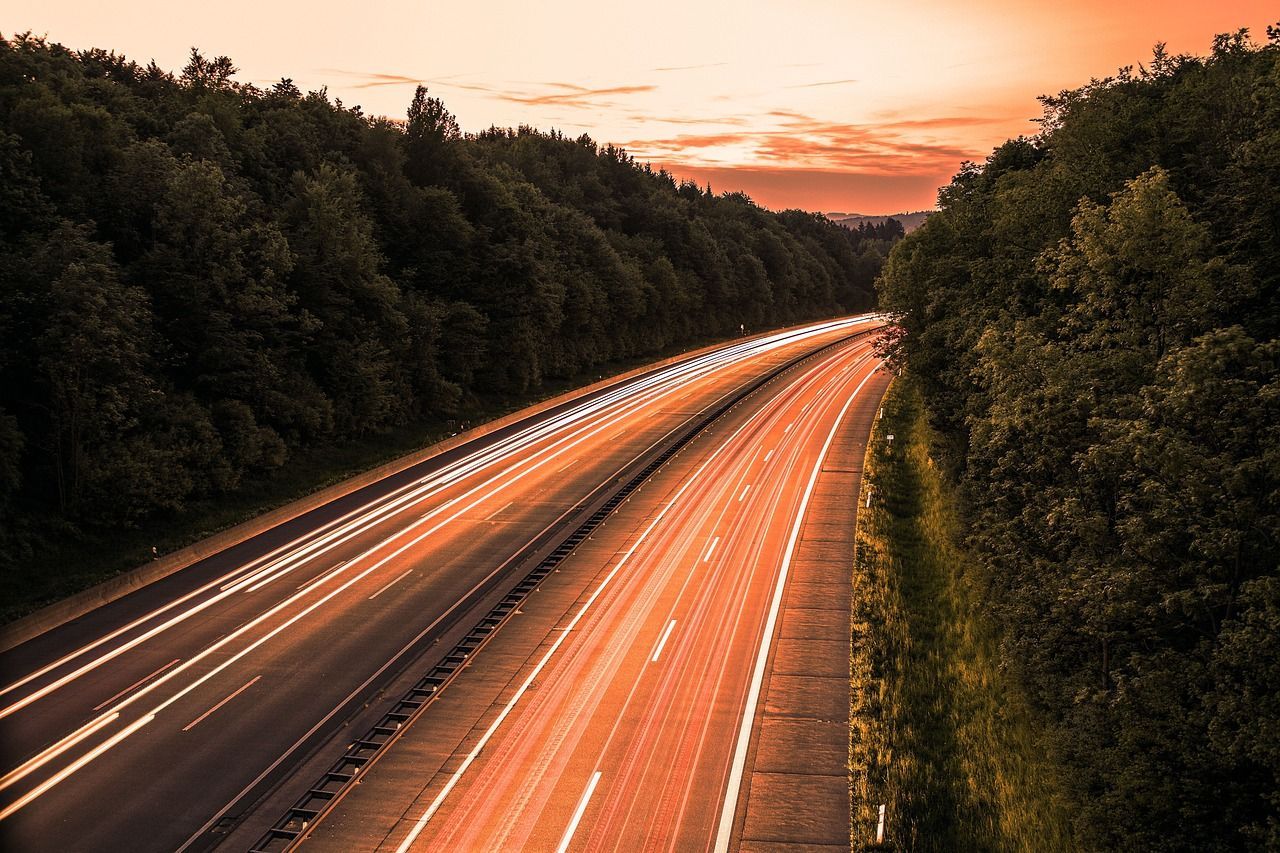 Een lange belichtingstijdopname van snelwegverkeer met gloeiende lichtsporen dat zich bij zonsondergang door een bos beweegt.