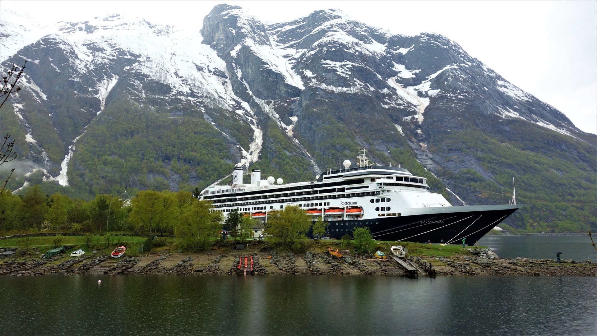 Een groot cruiseschip ligt aangemeerd aan een rotsachtige kustlijn voor een majestueus, met sneeuw bedekt berglandschap in een Noorse fjord.