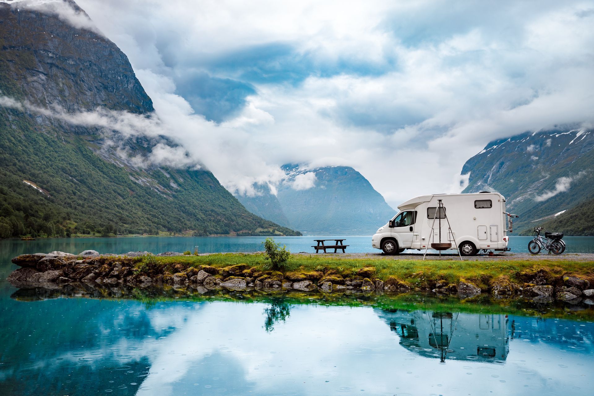 Een witte camper staat geparkeerd op een grasveldje aan de oever van een turquoise bergmeer, met weerspiegelingen in het water onder een bewolkte hemel.