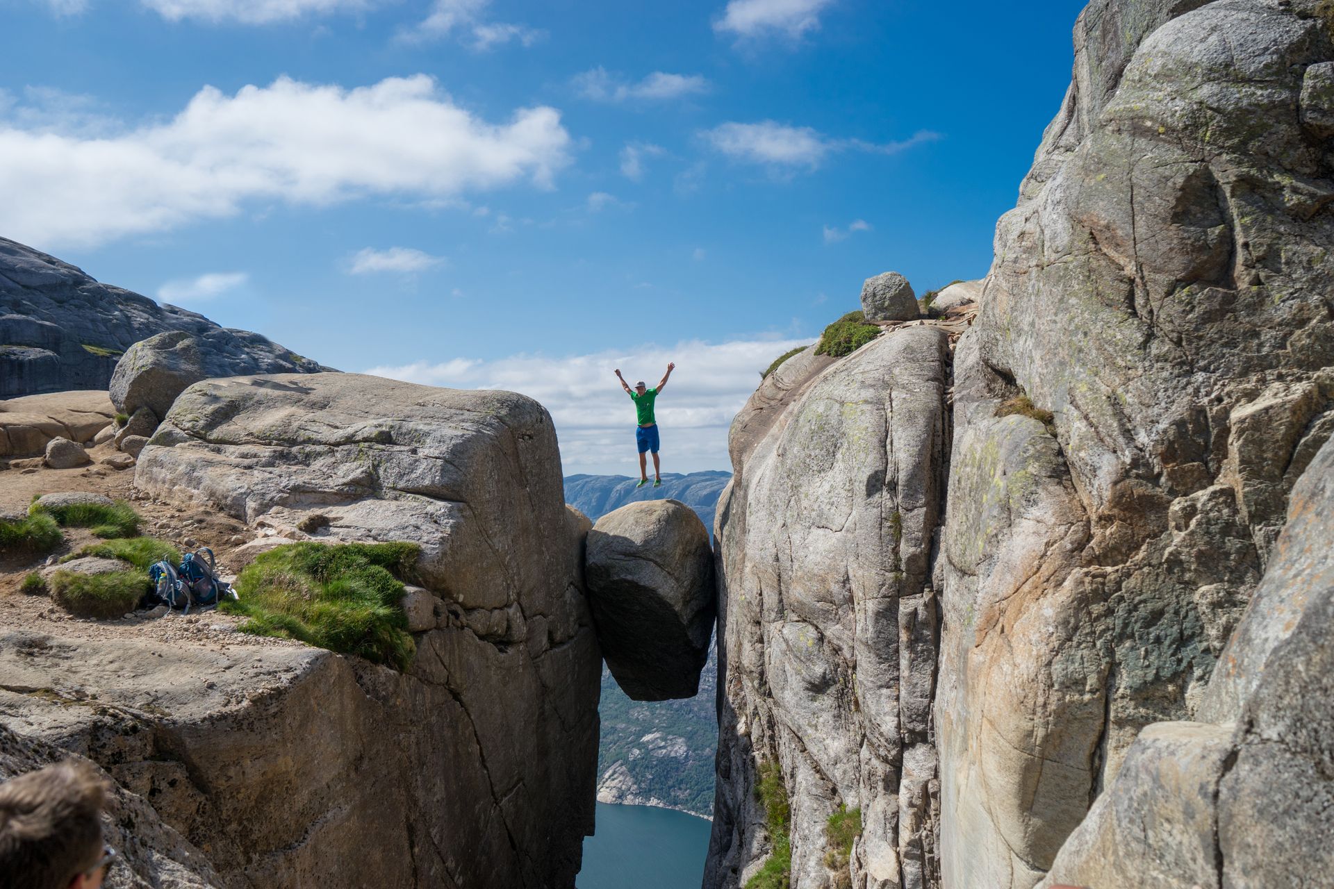 Met opgeheven armen staat een persoon op de Kjeragbolten-rots, die vastzit in een diepe bergspleet boven een diepblauwe fjord.