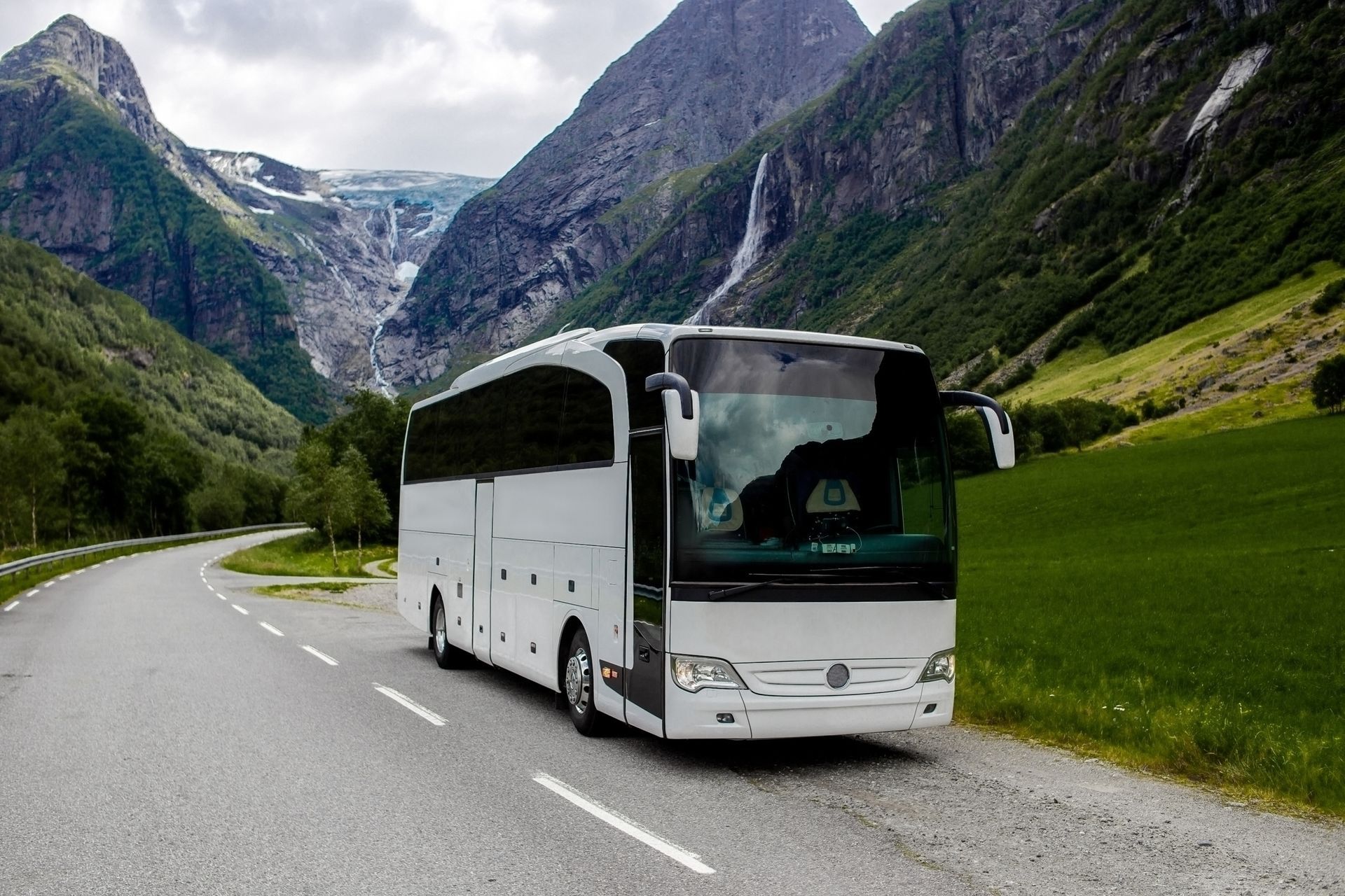 Een witte touringcar rijdt over een weg door een schilderachtige vallei, geflankeerd door steile, groene bergen en een waterval.