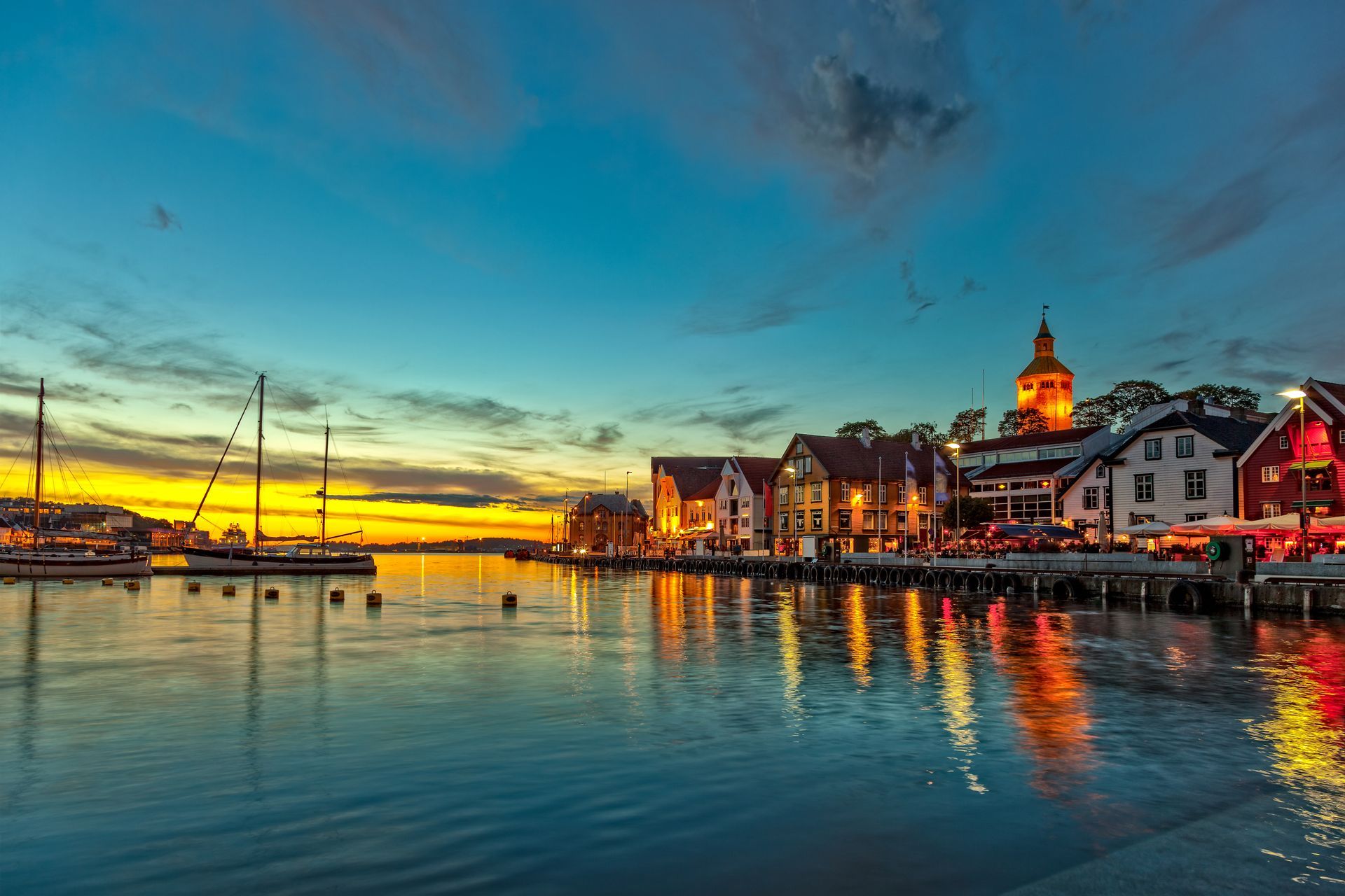 De haven van een kustplaatsje bij zonsondergang, met boten, historische gebouwen en warm goudkleurig licht dat weerkaatst op het kalme water.