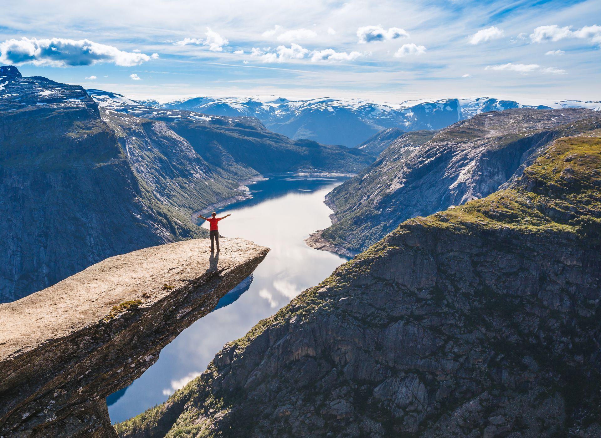 Een persoon in een rood shirt staat met uitgestrekte armen op Trolltunga, een rotsformatie die uitkijkt over een diep bergmeer.
