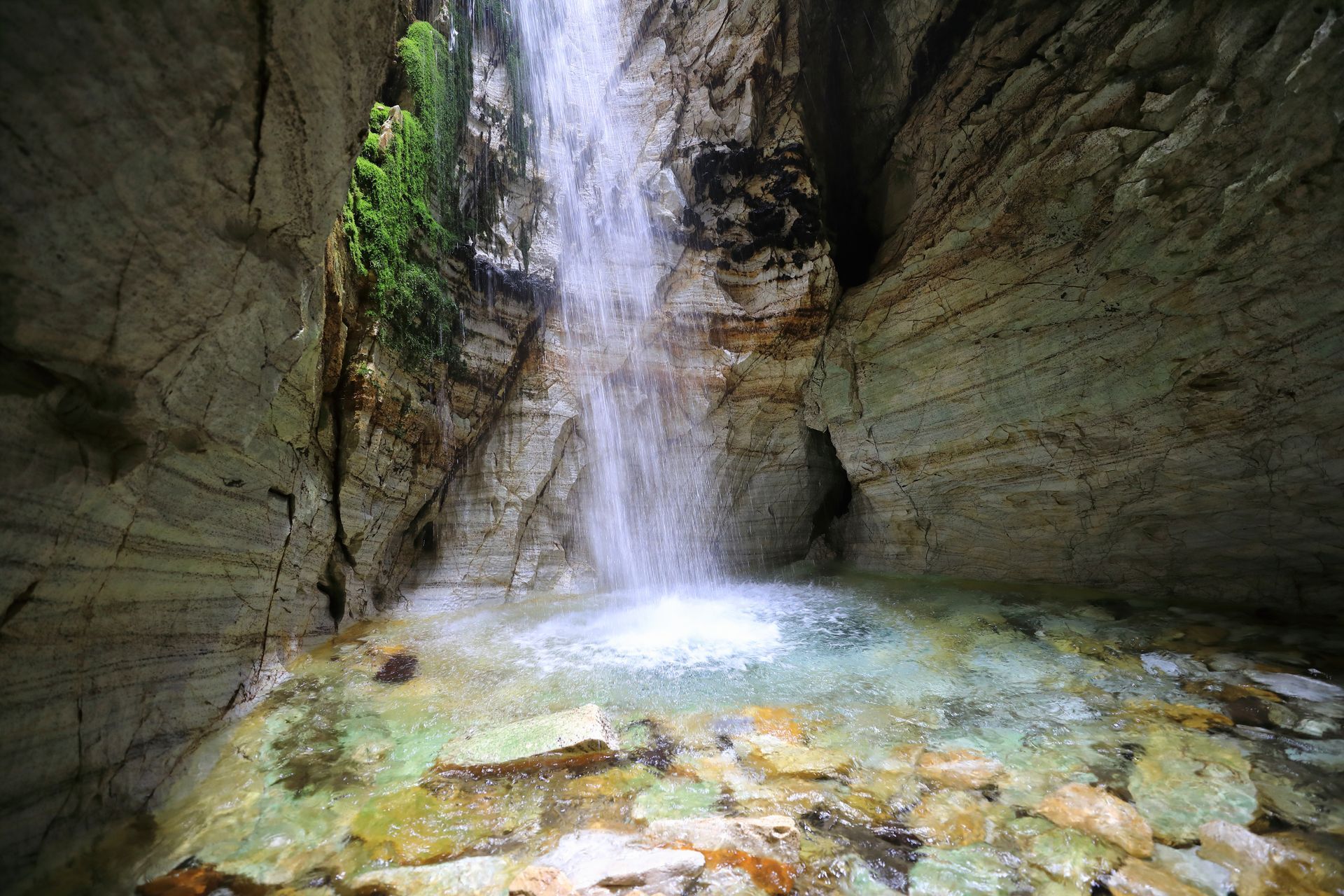 Een smalle waterval stort zich tussen de ruwe, rotsachtige canyonwanden naar beneden in een heldere poel gevuld met gladde, lichtgekleurde stenen.