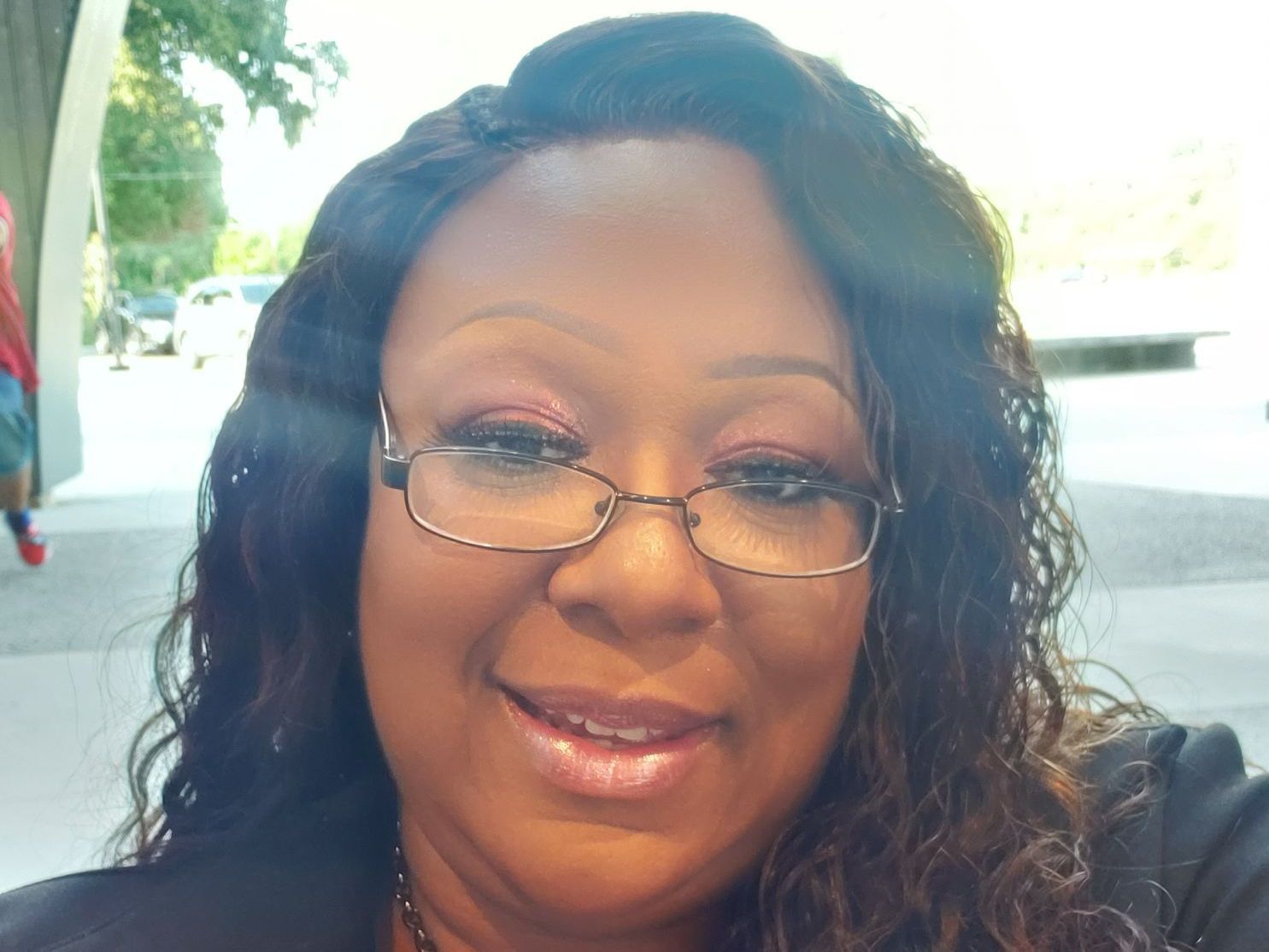Woman with glasses smiles at the camera outdoors. She has dark hair and makeup.