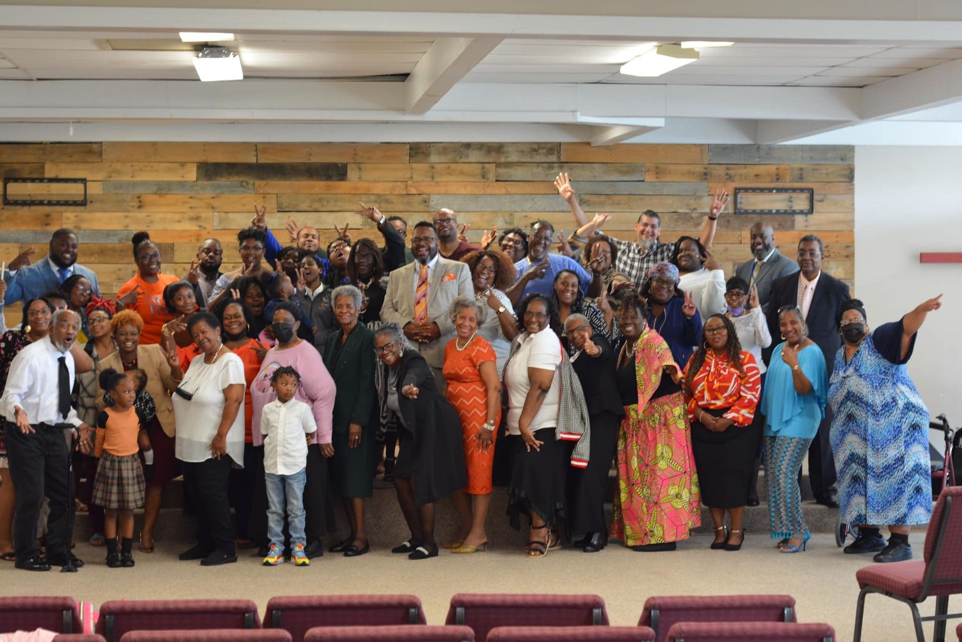 Group photo of diverse people indoors, many smiling and raising hands.