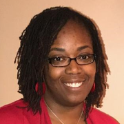 Woman with locs, glasses, and red earrings smiles, wearing a red blazer.