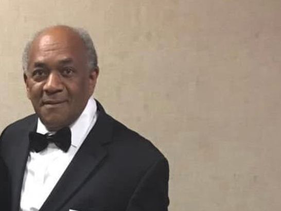 Older Black man in a tuxedo with a bow tie, smiling in front of a beige wall.