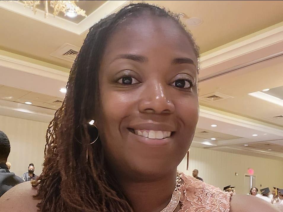 Woman with locs smiles, wearing a formal pink dress in a banquet hall.