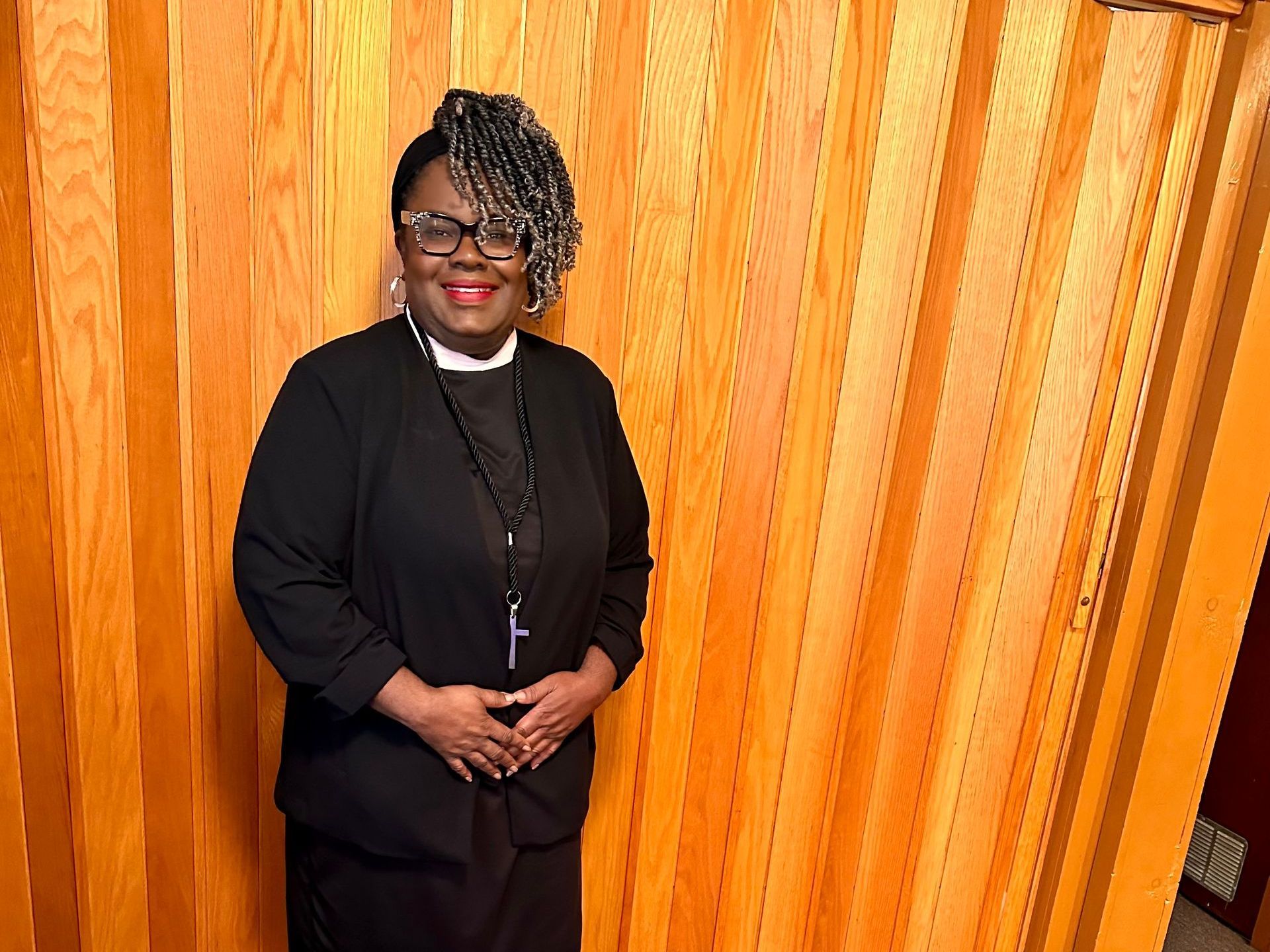 Black woman in clerical garb stands before a wood-paneled wall. She wears glasses, a cross necklace, and has gray hair.