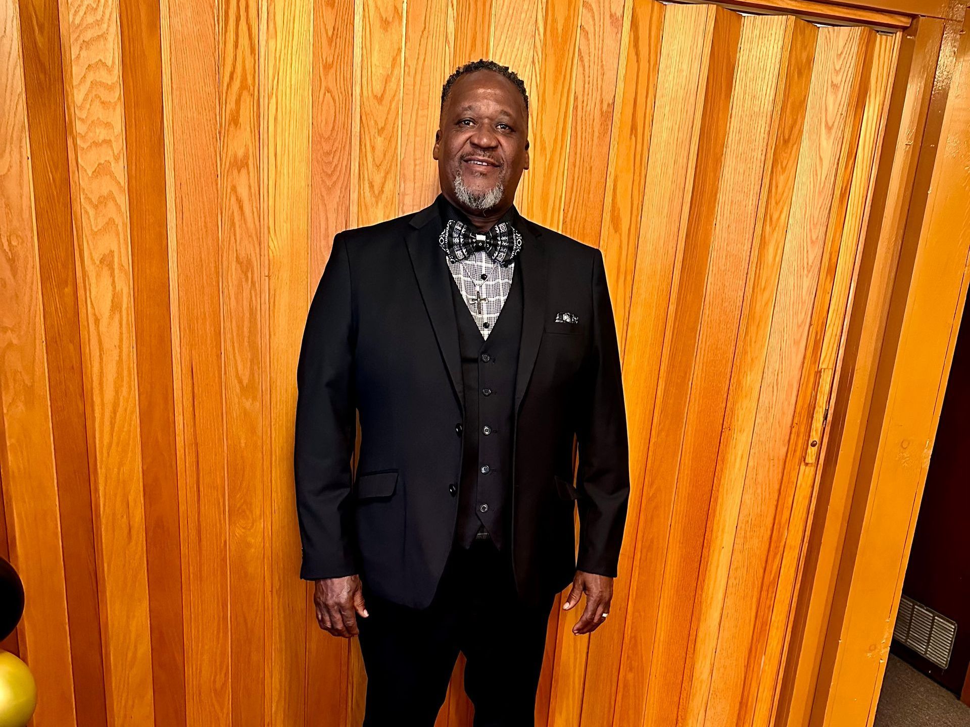 Man in black suit and bow tie stands against a wood-paneled wall.