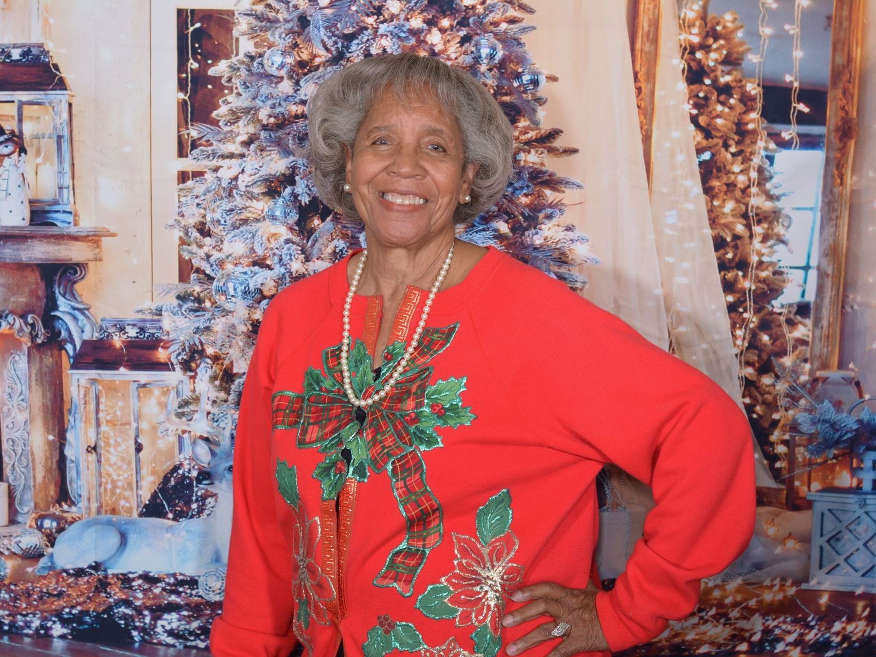 Older woman with gray hair in red Christmas sweater, smiling, in front of a holiday-themed backdrop.