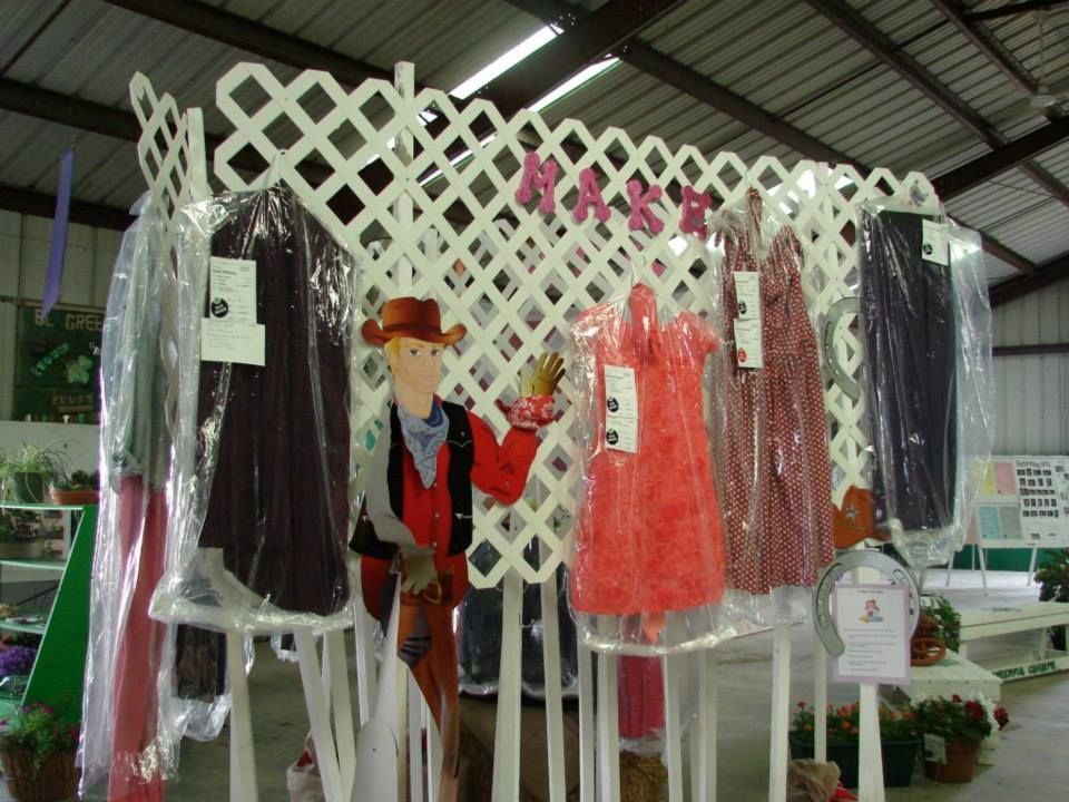 Garments in plastic covers hang on a white lattice display featuring a cardboard cutout of a person in a western outfit.