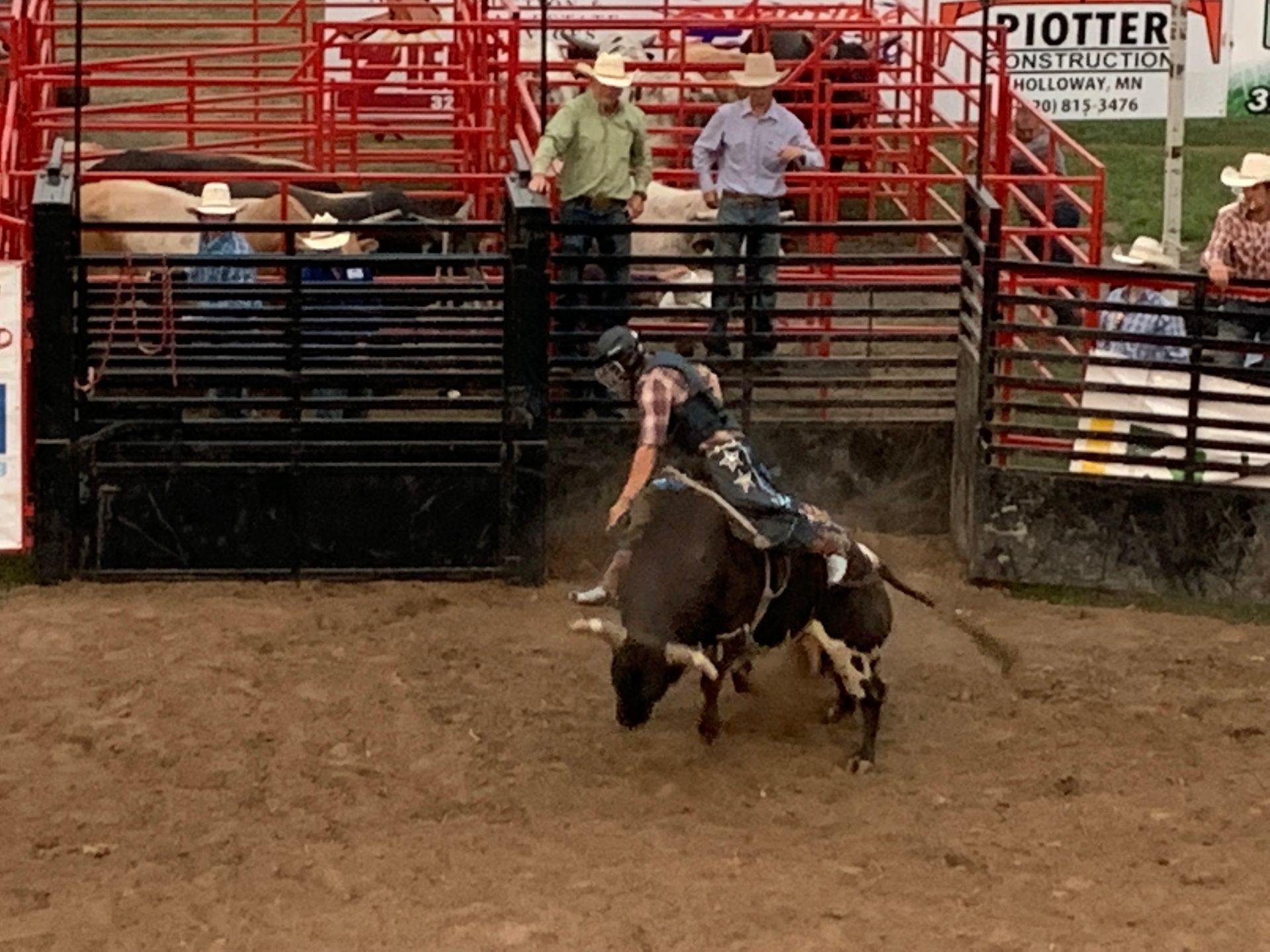 A bull rider in a plaid shirt rides a dark bull in an outdoor dirt arena, surrounded by metal fencing and spectators.