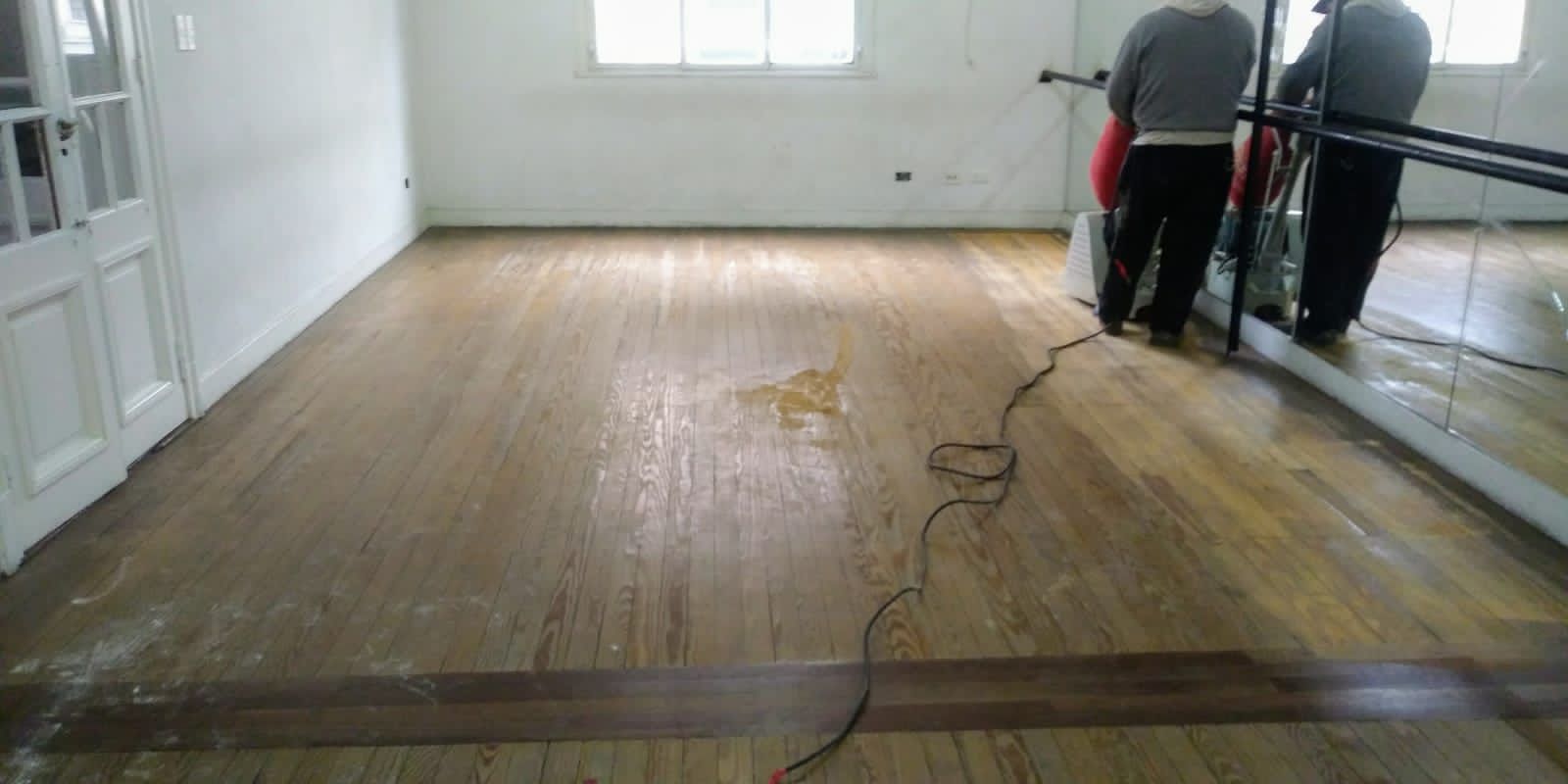 Dos trabajadores lijando un piso de madera en una habitación luminosa y vacía.