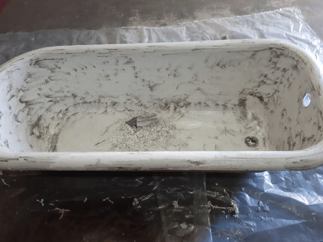Bañera blanca sucia con manchas y marcas de rozaduras, colocada sobre una superficie oscura bajo una lámina de plástico.
