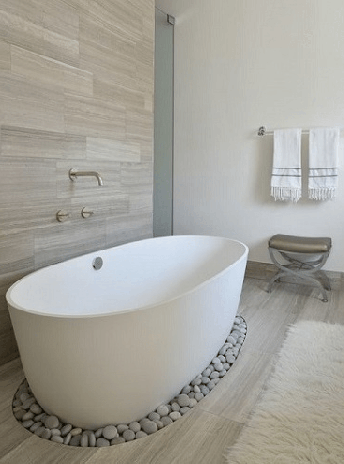 Baño moderno con bañera blanca independiente, borde de guijarros, toallero y pared de piedra texturizada.
