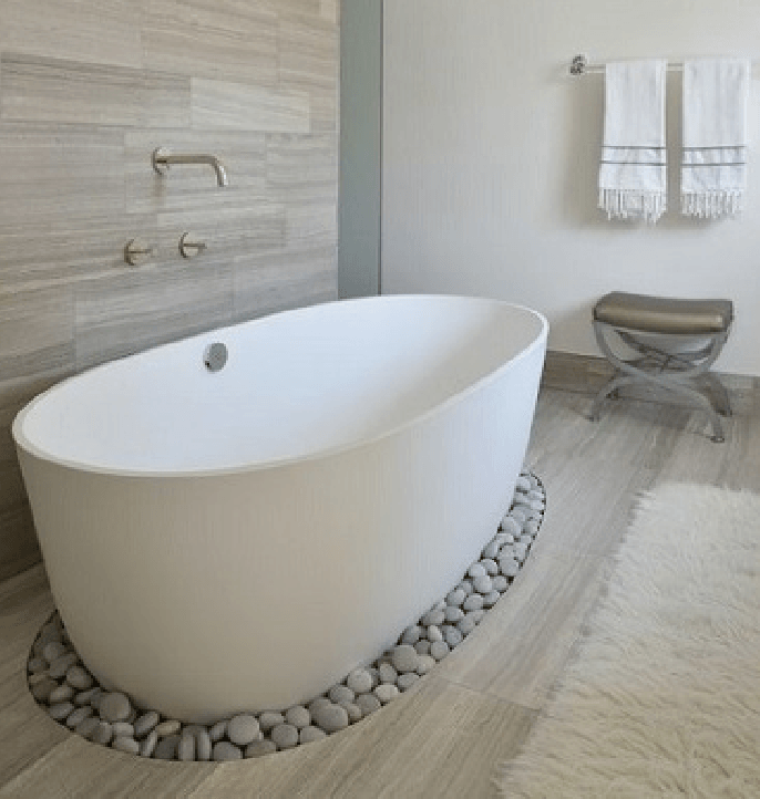 Baño moderno con bañera blanca independiente, base de guijarros y taburete cubierto con toalla.
