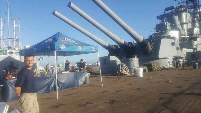 Naval guns on a ship deck beside a blue tent and a man standing outdoors under a clear sky
