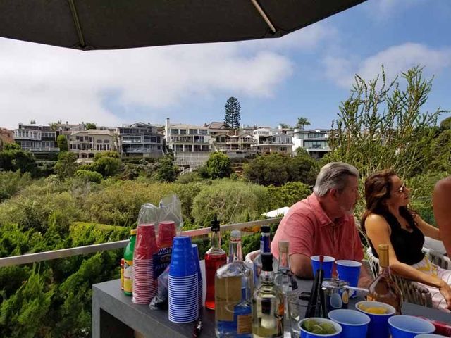 Outdoor patio with drinks on a table, overlooking greenery and white houses under a bright sky