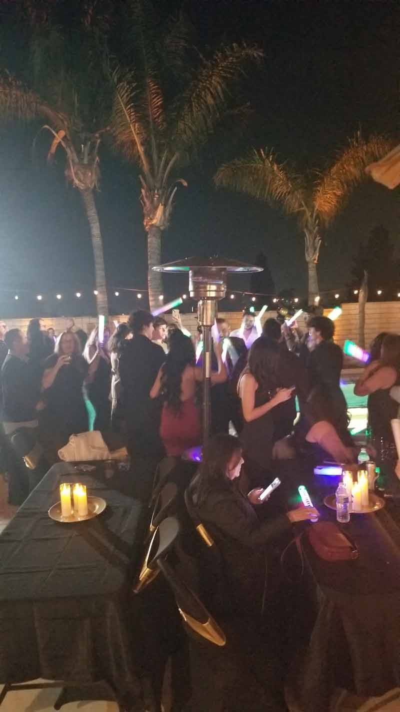 Outdoor evening party with guests seated at tables under palm trees, lit by warm candles and colorful lights