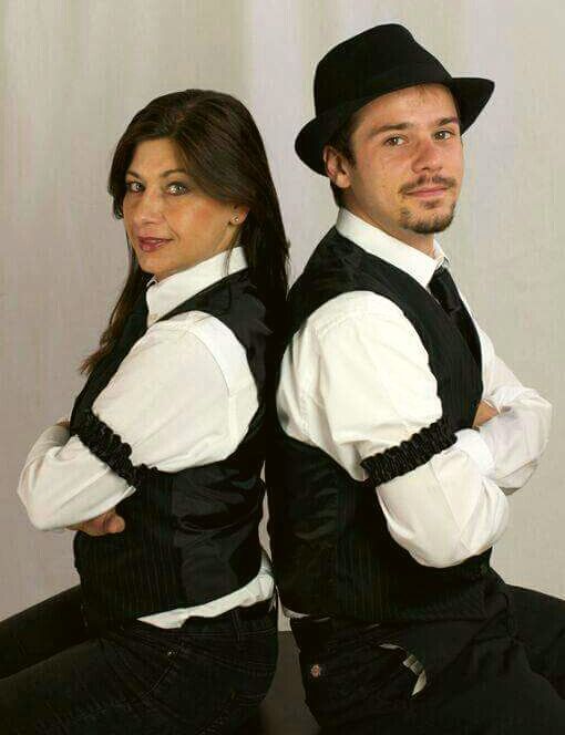 Two people in black vests and hats sit back-to-back, posing with crossed arms against a plain background.