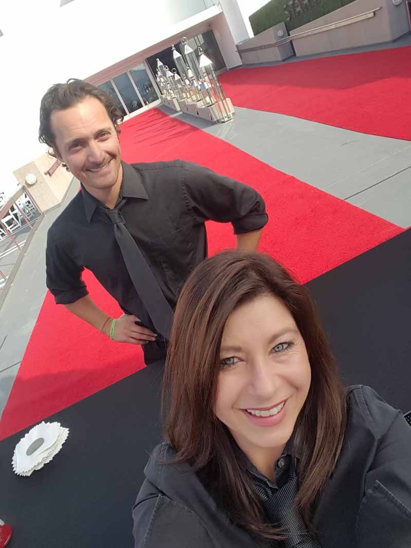 Two people smiling on a red-carpet walkway at an outdoor event.