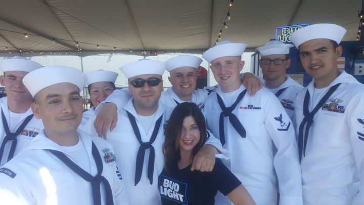 Group of sailors in white uniforms posing with a woman in a black shirt under a covered walkway.