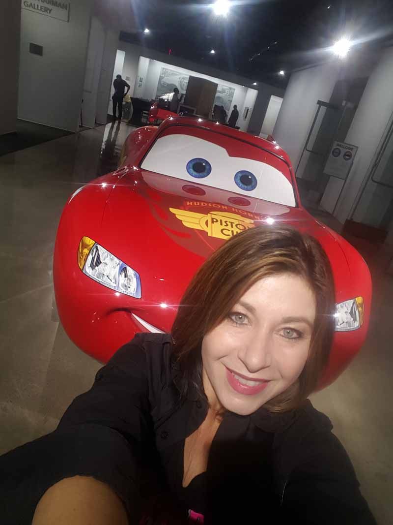 Woman taking a selfie with a red Lightning McQueen car in a hallway.