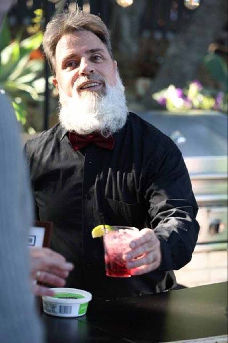 Man holding a drink beside a table outdoors, with a small tub in the foreground and blurred cars behind