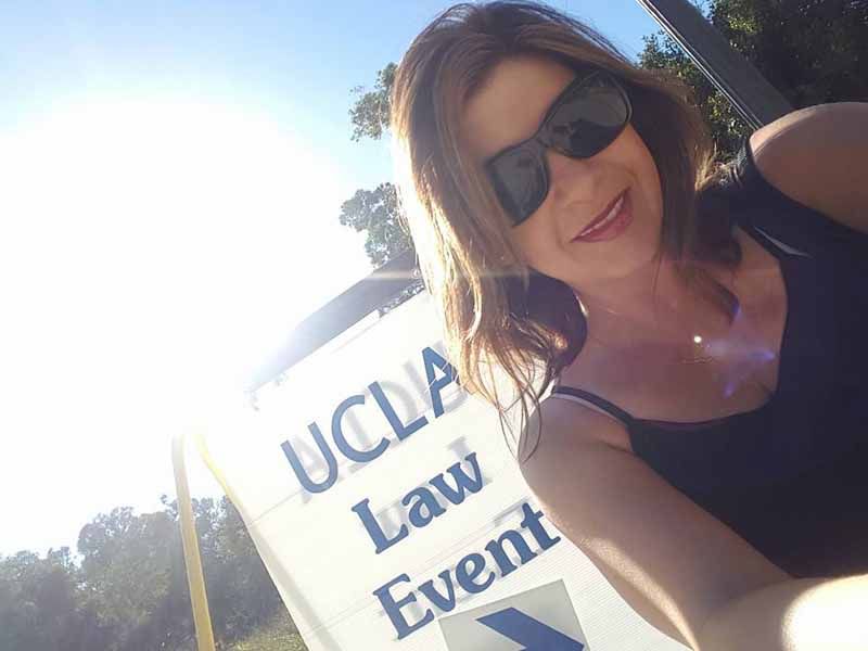 Woman in sunglasses posing beside a UCLA Law Event sign outdoors in bright sunlight