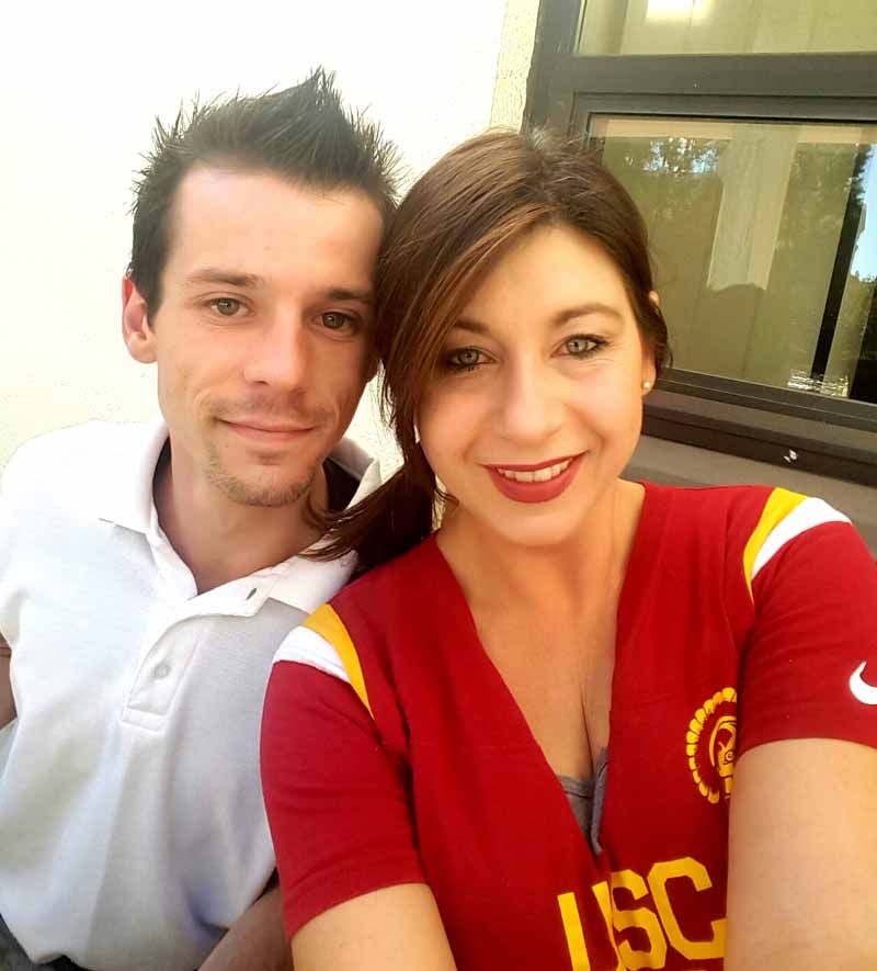 Two smiling people taking a selfie, one in a white shirt and one in a red USC shirt, indoors by a window