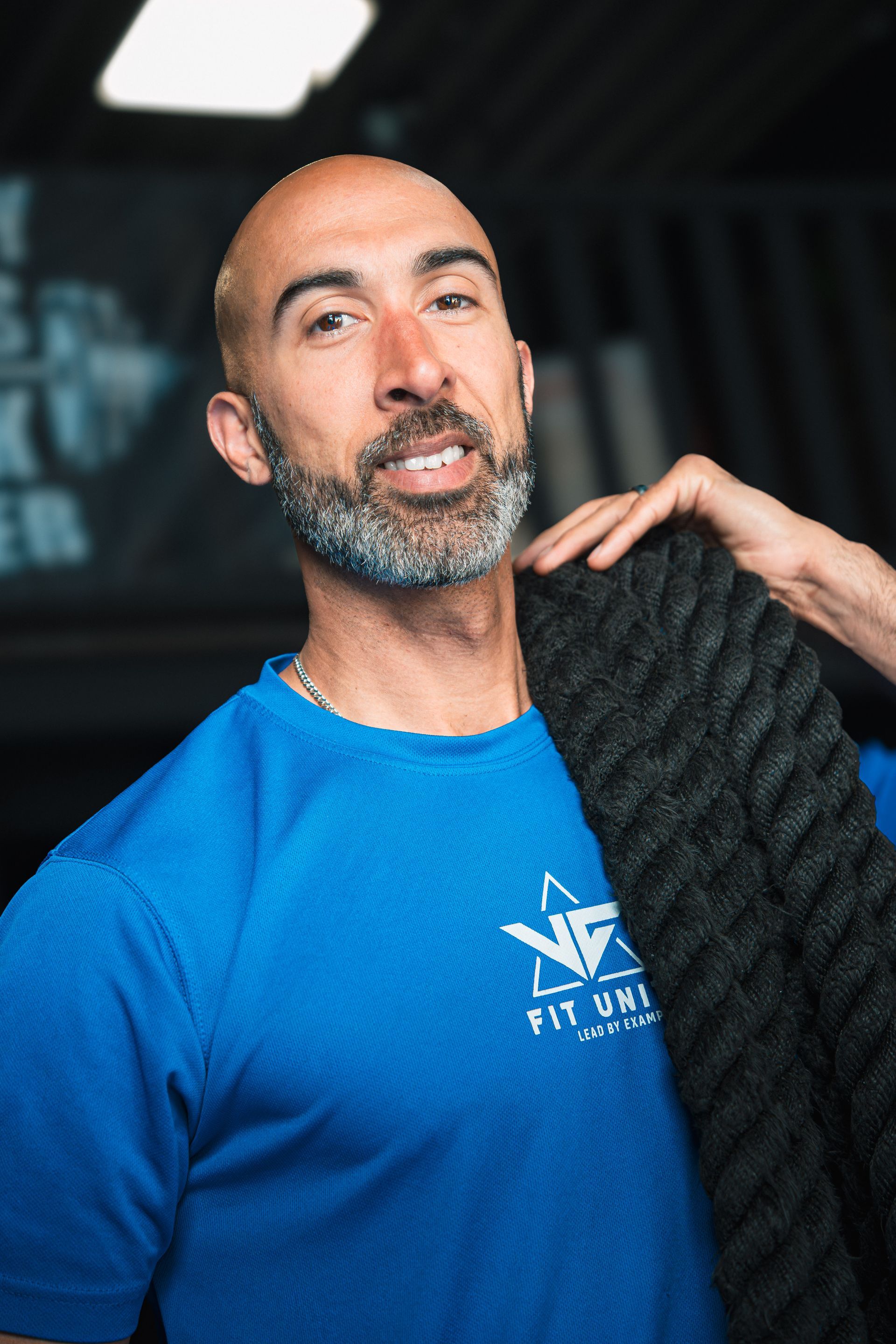 Een man in een blauw T-shirt, met een zwarte, gestructureerde jas over zijn schouder, staat binnen te glimlachen.