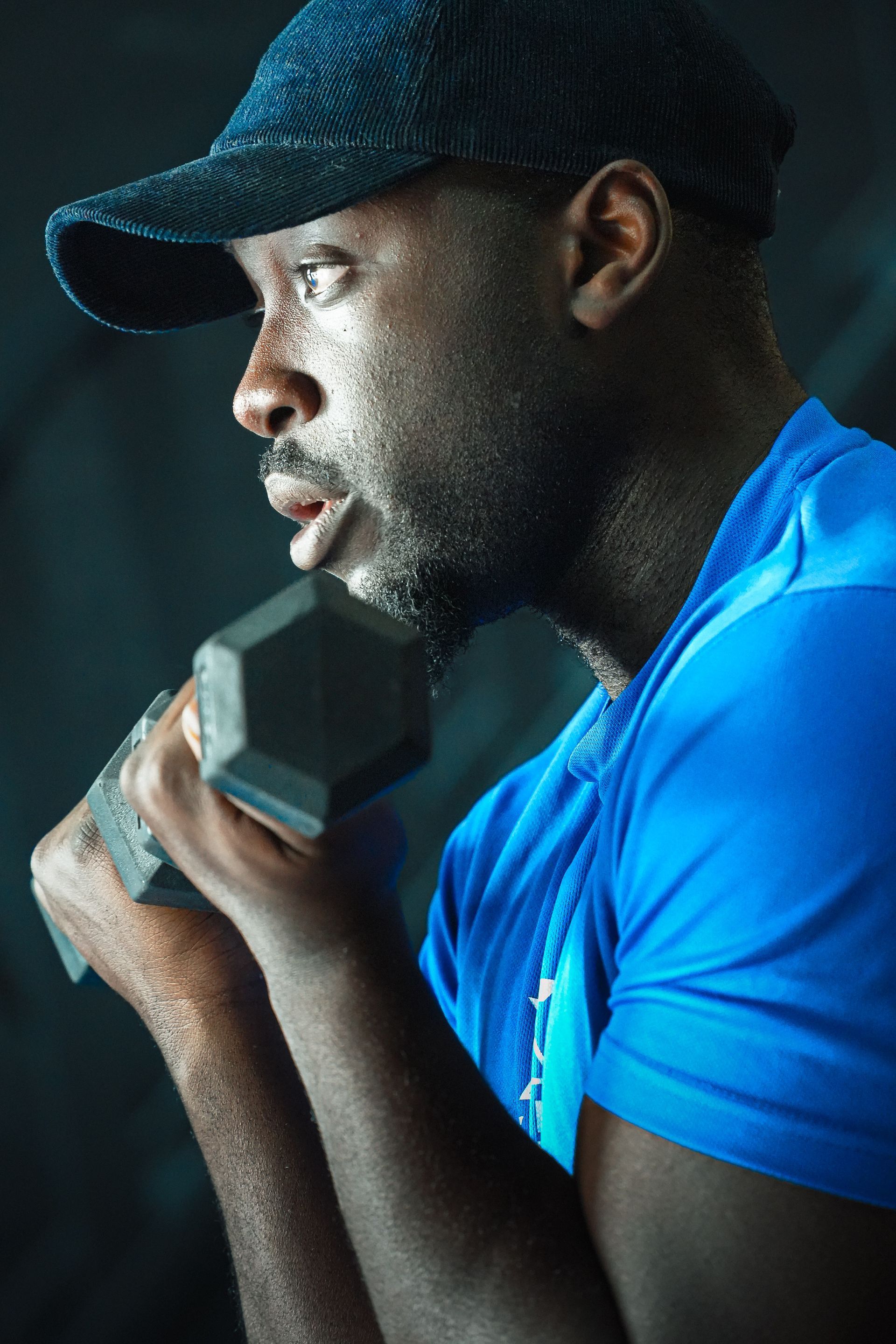 Persoon in blauw shirt en pet tilt een halter op in een sportschool, zijprofiel.
