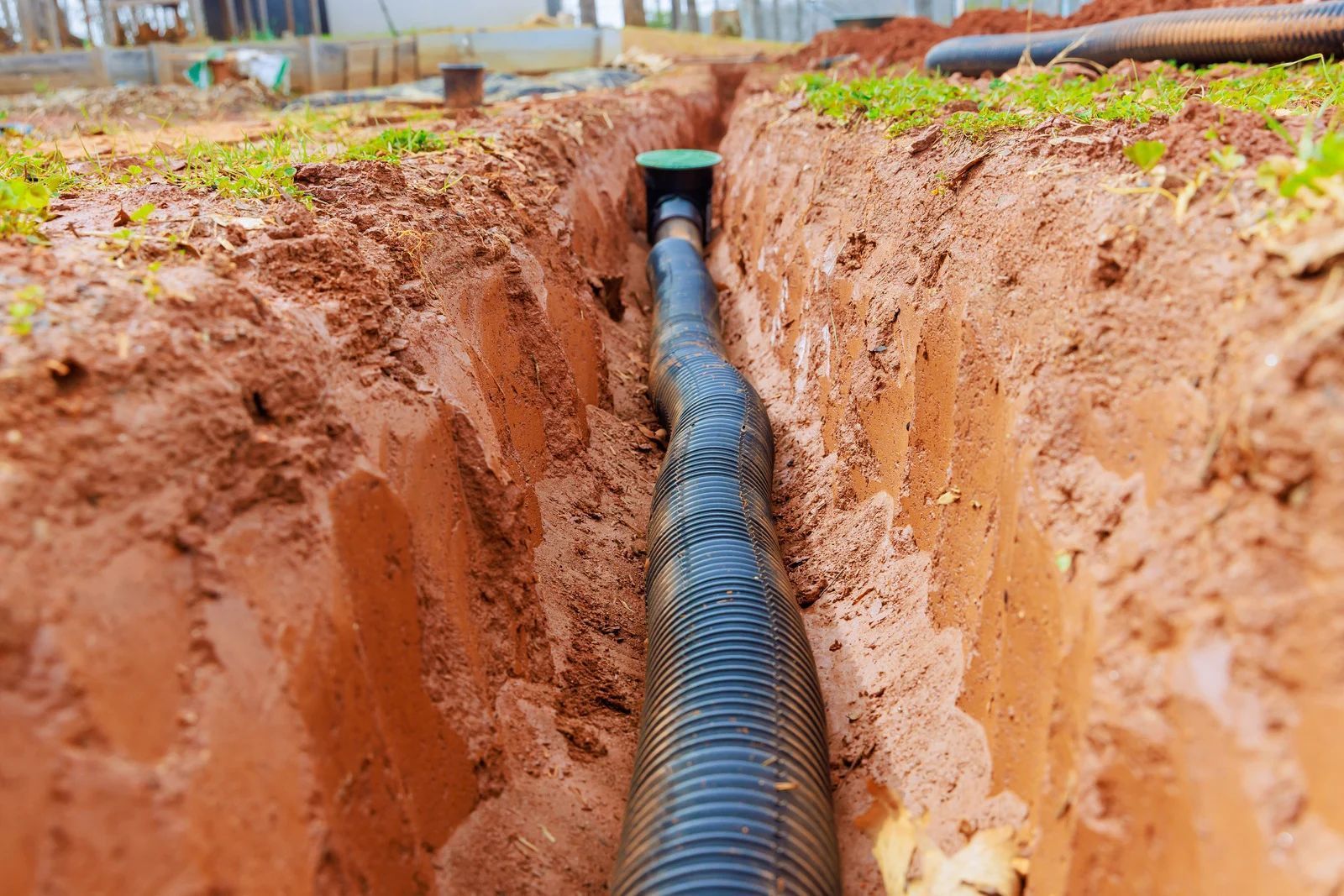 A black, flexible, corrugated drainage pipe laid inside a freshly dug trench in red soil, leading to a small green cover.