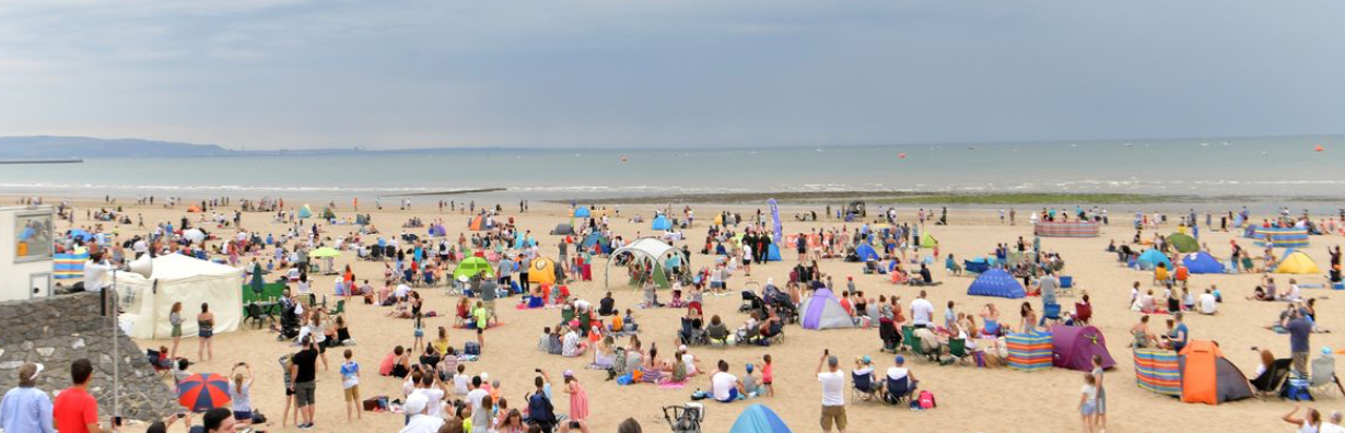 Crowded beach with people relaxing, tents, and a gray sky. The ocean stretches in the background.