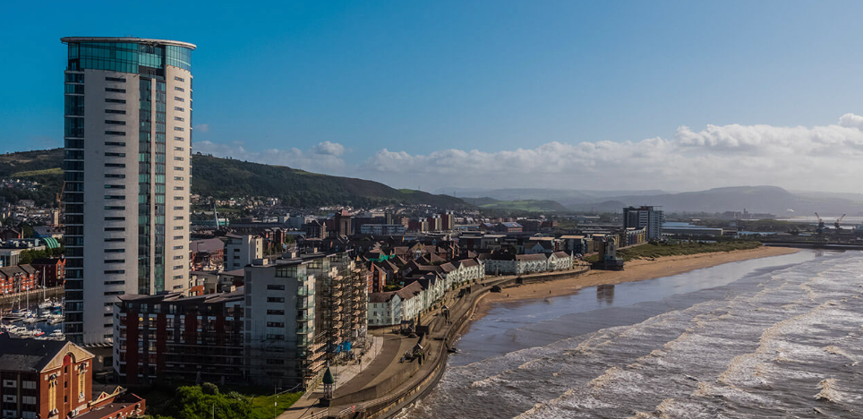 Coastal cityscape with a tall skyscraper, beach, and mountains under a bright blue sky.