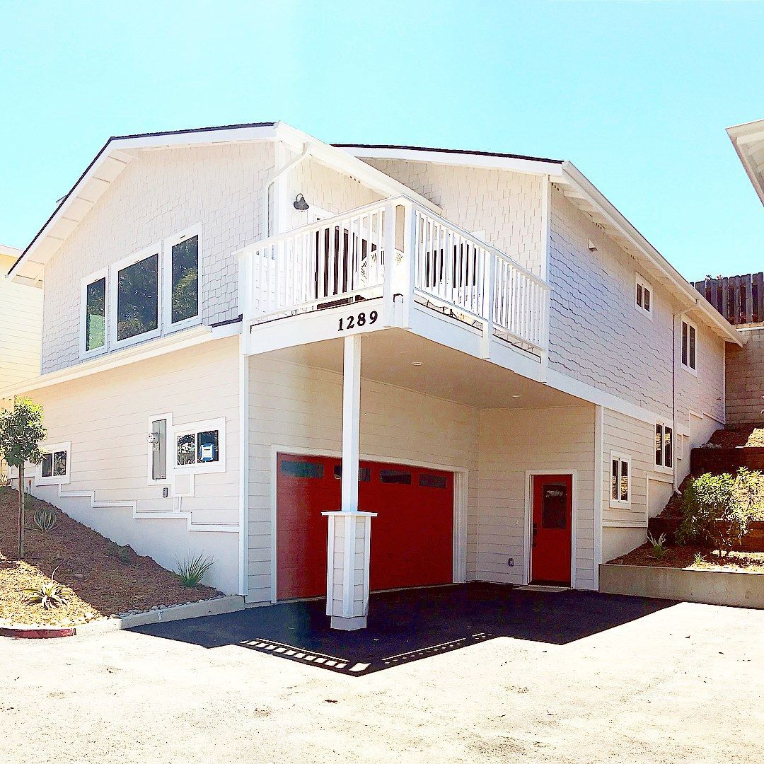 Two-story white house with a red garage door, balcony, and front door. Blue sky overhead.