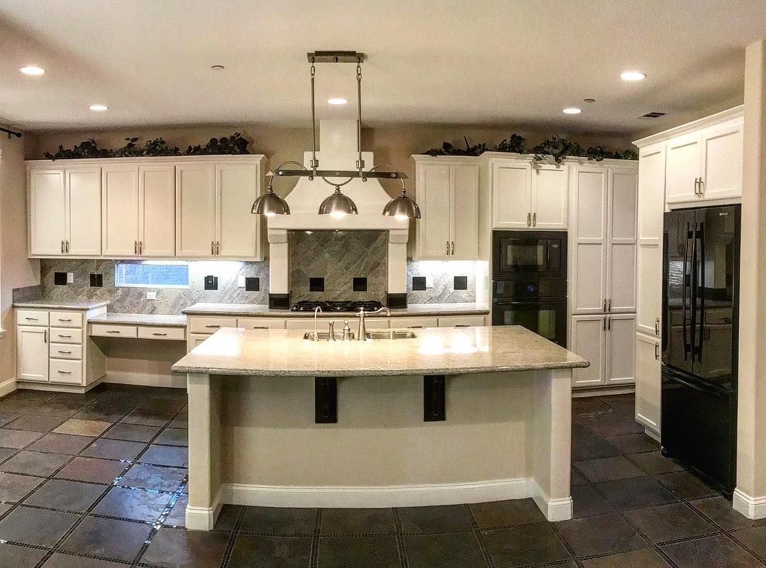 Cream-colored kitchen with island and black appliances, granite countertops, and dark floor tiles.