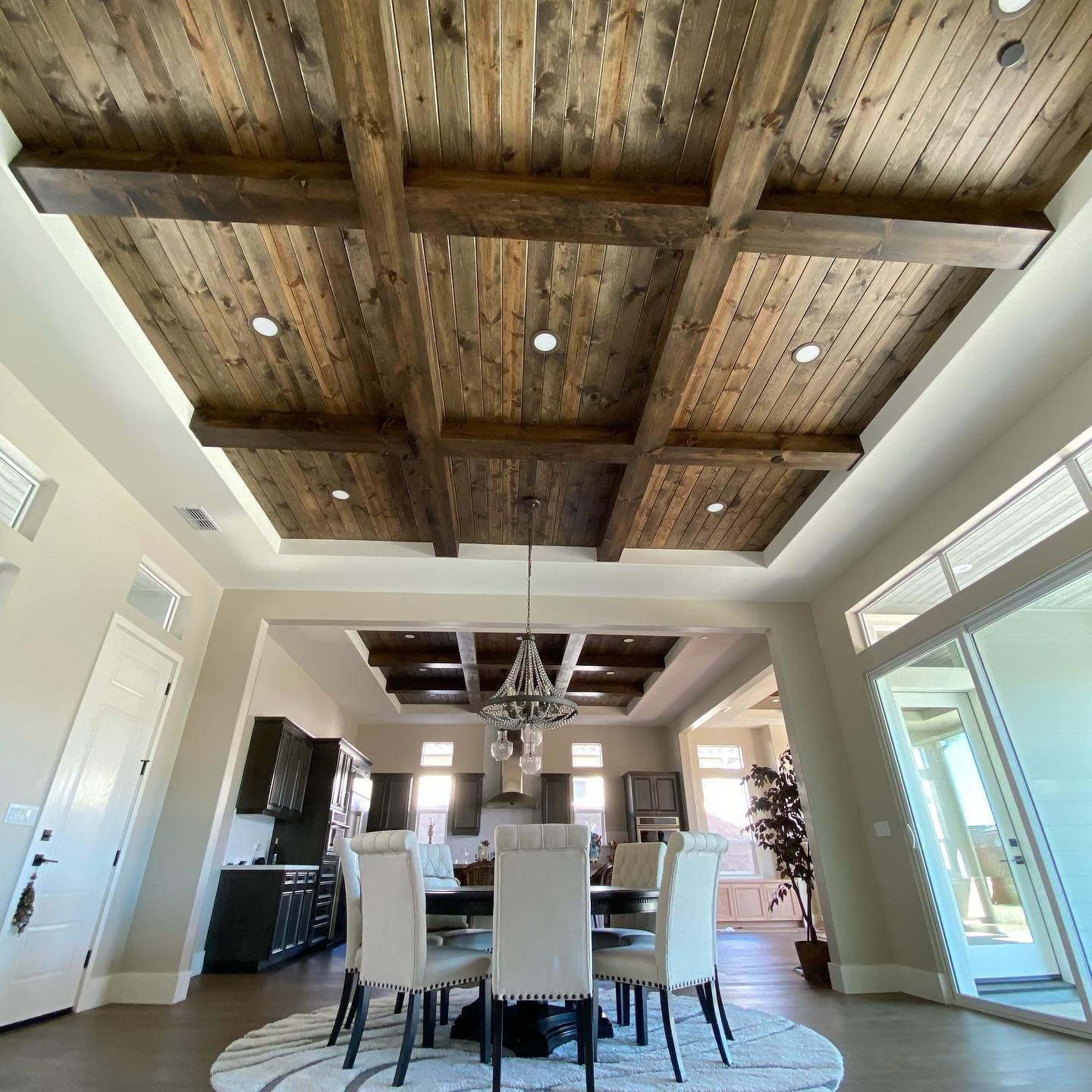 Dining room with wooden ceiling beams, chandelier, table, chairs, and kitchen in the background.