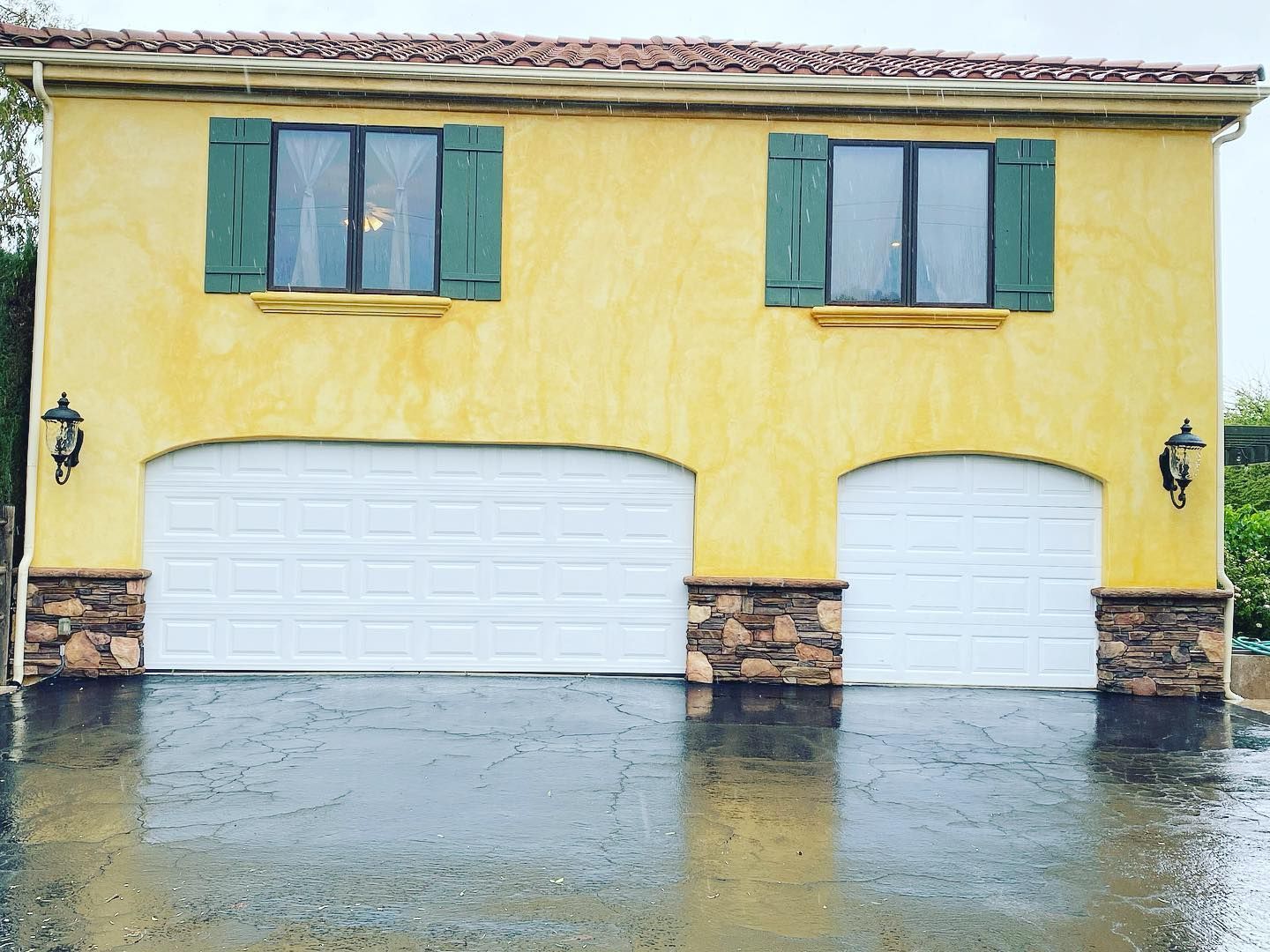 Yellow stucco garage with two white doors, green shutters, and brown stone accents, rain falling.