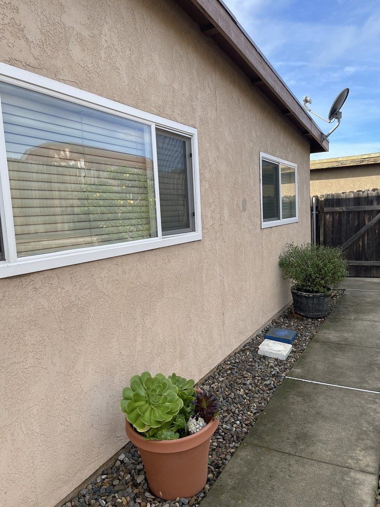Tan stucco building with two windows, a satellite dish, and potted plants.