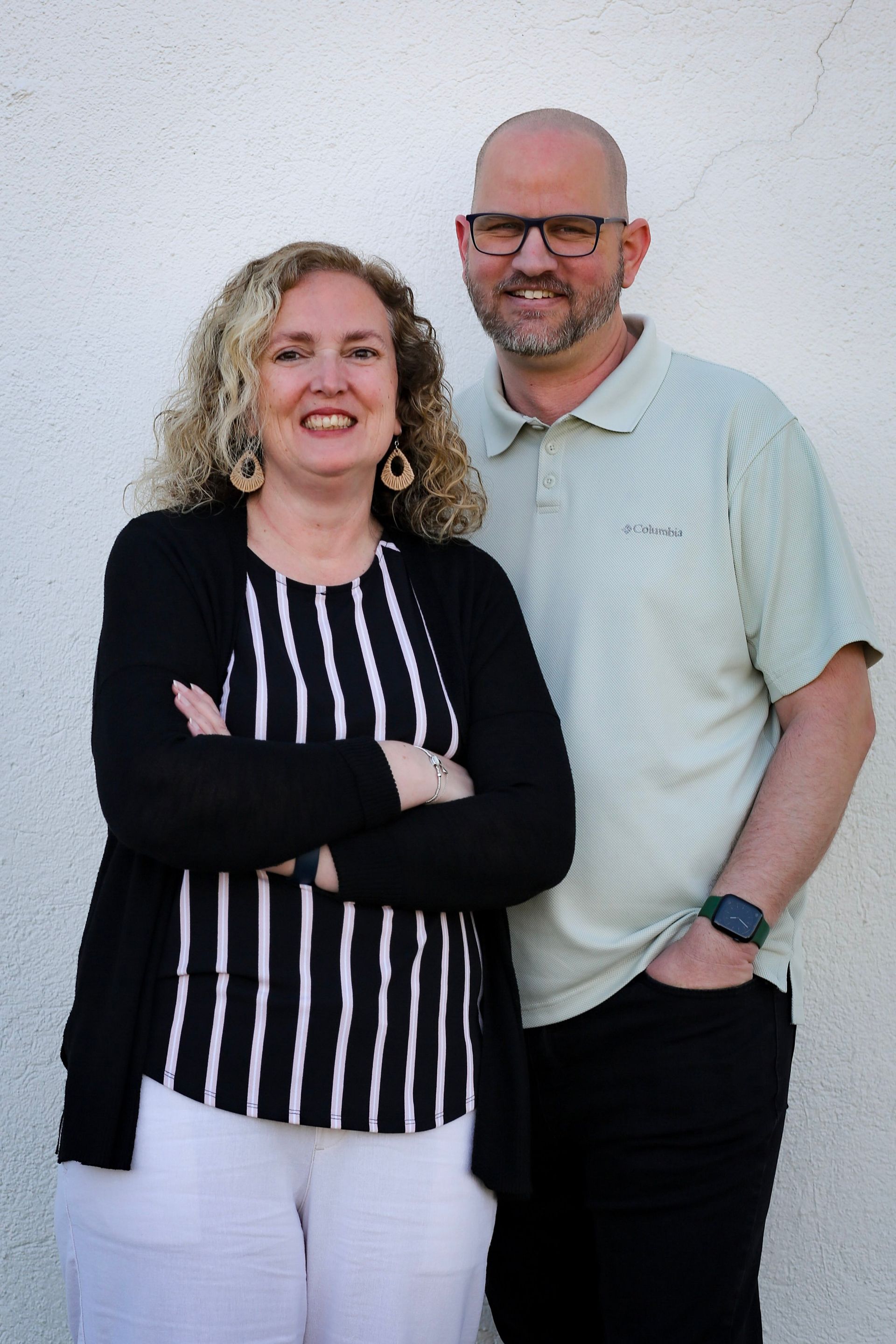 Two smiling people standing against a white wall, one with arms crossed, both in casual clothes