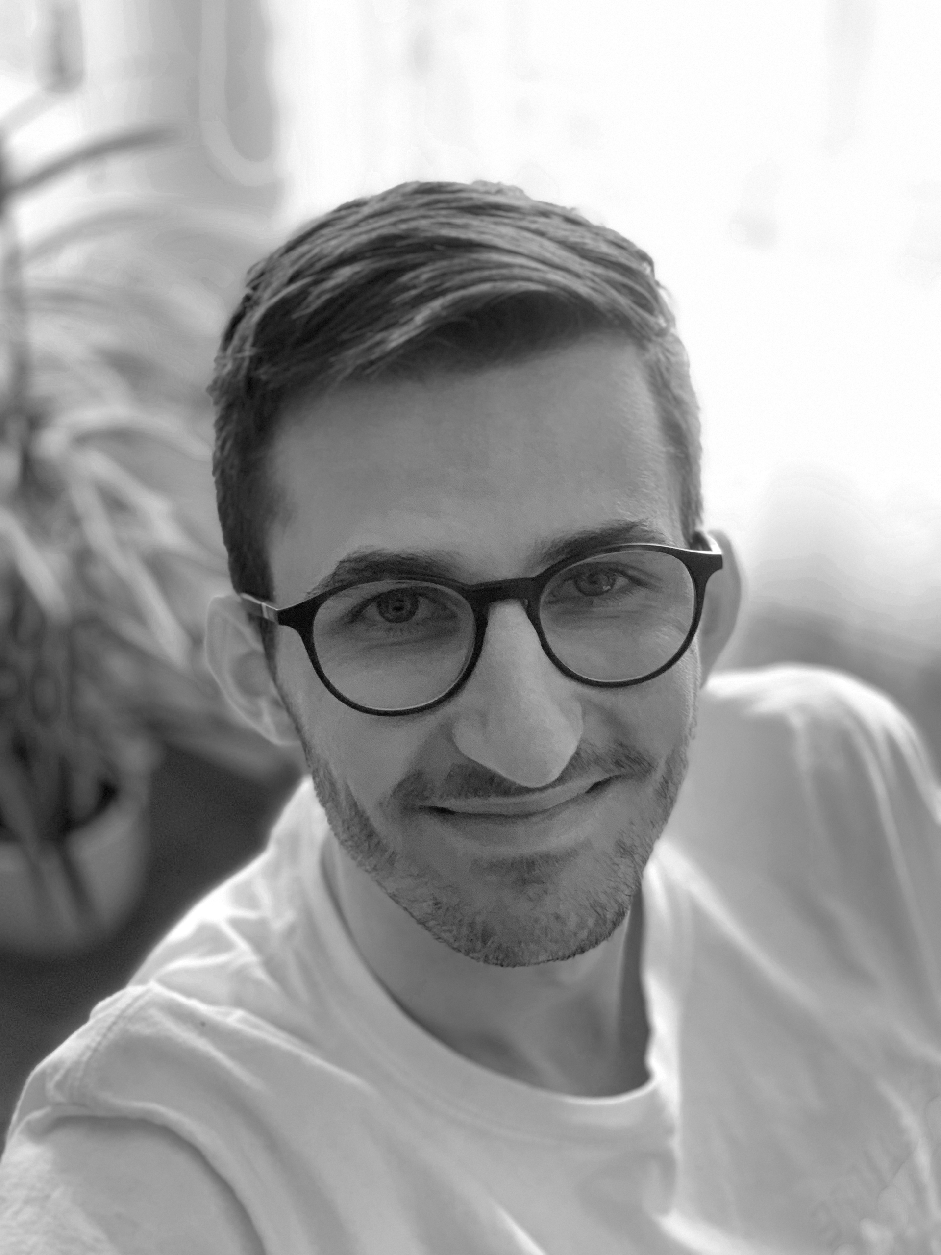 Black-and-white selfie of a smiling person wearing round glasses and a white shirt indoors